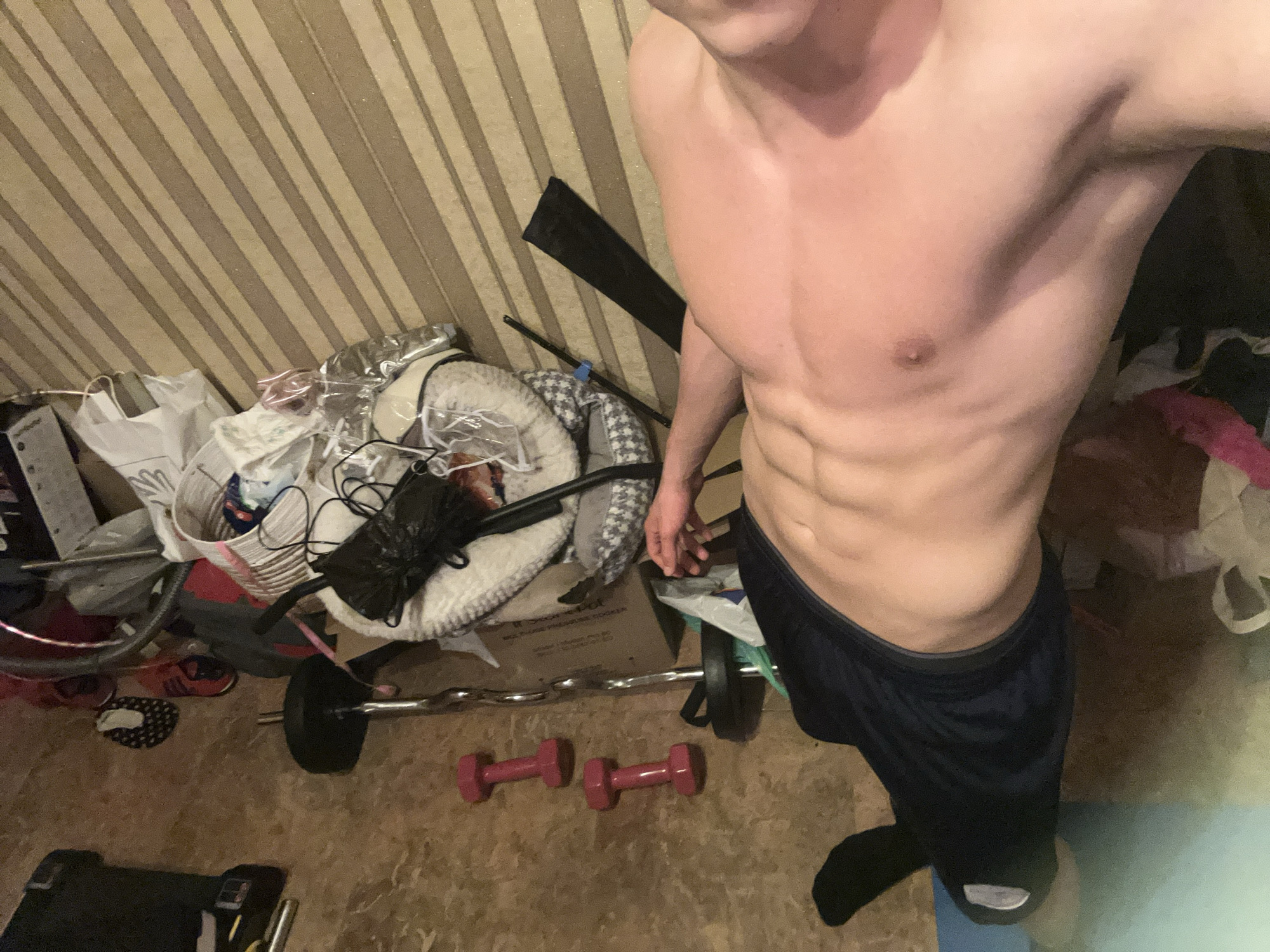 The image depicts a shirtless man with visible abdominal muscles standing in a cluttered room. The room's walls are covered in striped wallpaper. Scattered around the floor are various items, including a barbell, two small pink dumbbells, bags, a pet bed covered in plastic, a black mask, and sports equipment. The exact location within Burgas, Bulgaria, is not discernible from the image.
