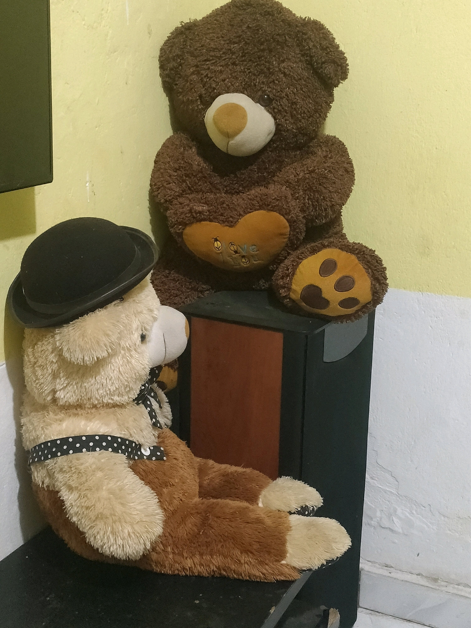 Two teddy bears are positioned on a dark surface against a yellow and white wall. The bear in the foreground is light brown and cream-colored, wearing a black bowler hat and a black and white polka dot ribbon around its neck. It is sitting and facing to the left, with its head slightly tilted. The bear behind it is dark brown, sitting upright, and facing forward. It has a heart-shaped patch on its chest that appears to have stitching on it. The scene is likely indoors, as indicated by the wall and the presence of what appears to be a speaker or electronic device below the dark brown bear. The image does not contain sufficient visual information to determine its specific location within the city of Pekanbaru, Indonesia.