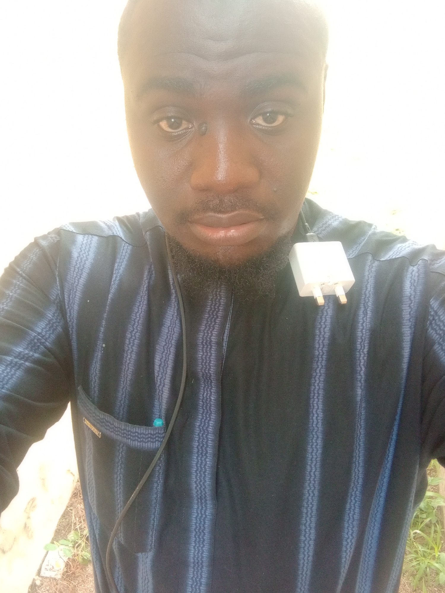 The media file is a medium close-up selfie of a dark-skinned man, likely taken outdoors in Jalingo, Nigeria. The man is wearing a dark blue, vertically striped traditional garment with a V-neck. A white electrical plug, possibly part of a phone charger, is attached to the garment near his collarbone, with its cord hanging down. He has a short beard and mustache, and his eyes are looking directly at the camera. There is a faint mole above his right eye. The background is a bright, washed-out white, suggesting it might be daytime with strong sunlight or a bright, evenly lit wall. The overall lighting suggests it is daytime. The man's expression is neutral. A small, rectangular label with faint text is visible on the left chest pocket of his garment. A small turquoise object is visible in the pocket. The scene appears casual, with some out-of-focus greenery and earthy ground visible at the bottom edges of the frame.