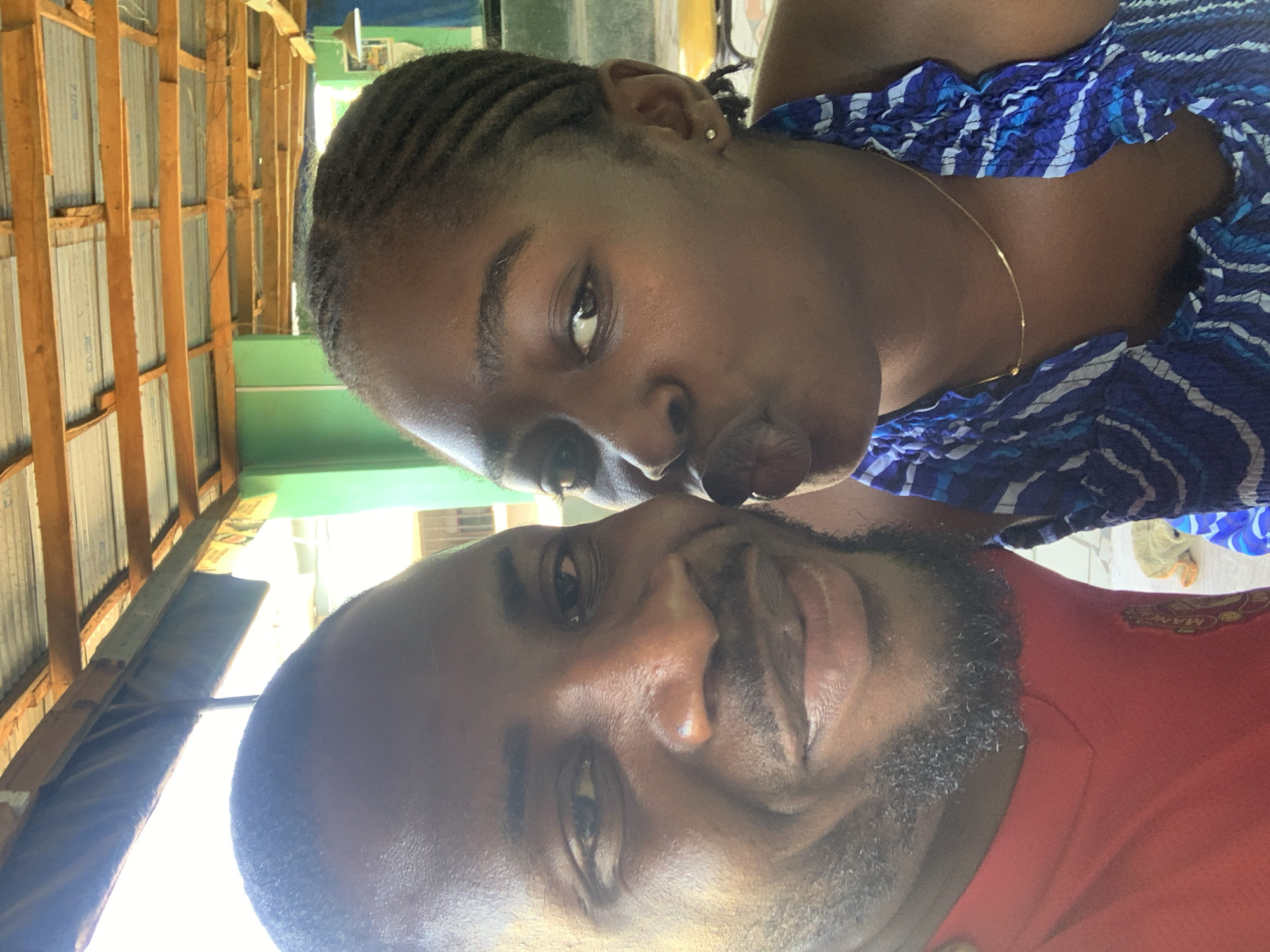 Two people are close together in a selfie-style photograph. The person at the top has dark skin, braided hair, is wearing a blue and white patterned top, and has a thin gold necklace. The person at the bottom has dark skin, a beard, and is wearing a red shirt. The background shows a wooden structure with corrugated metal sheets and a green section. There is nothing in the photo to specifically indicate the location is New Stadium, Nigeria.