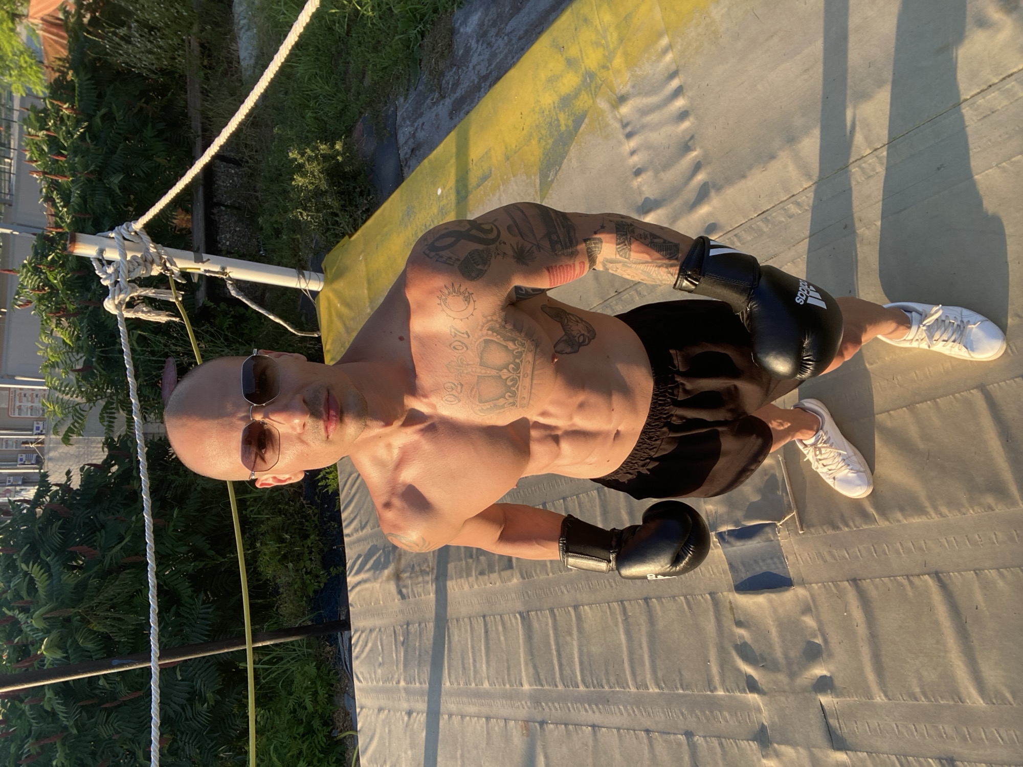 An athletic, bald man with sunglasses stands in a boxing ring. He has a muscular physique and is covered in tattoos on his arms and chest. He is wearing black boxing shorts and black boxing gloves, one on each hand. The ring appears to be outdoors, with trees and some buildings visible in the background. The ground of the ring is a padded mat with yellow accents. The man is looking directly at the camera. The scene is lit by the golden light of what appears to be sunset. The image was taken in the city of Pleven, Bulgaria.