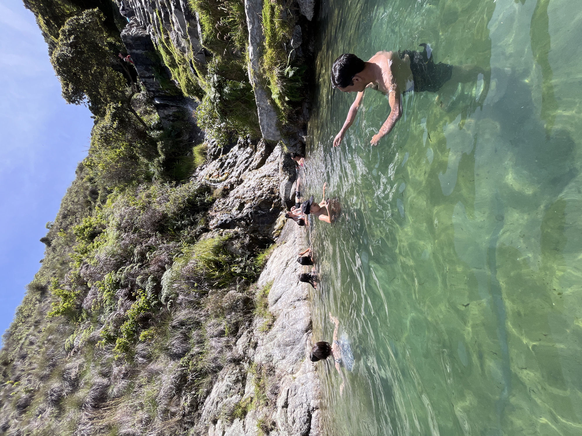 The image shows a natural pool of clear green water situated within a rocky, vegetated mountainous terrain. Several people are swimming and interacting in the water. In the foreground, a shirtless man with dark hair is partially submerged, extending his right arm forward. To his left, a person wearing a patterned swimsuit is visible from the back. Further into the pool, other individuals are seen swimming, with some with their heads above water. The background features steep, rocky inclines covered with lush green vegetation, grasses, and some trees. A stone staircase and a small waterfall are discernible on the right side of the frame, indicating a developed access point to the natural feature. The sky above is bright blue, suggesting a sunny day. The location is identified as a natural pool in Mucurubá, Venezuela.