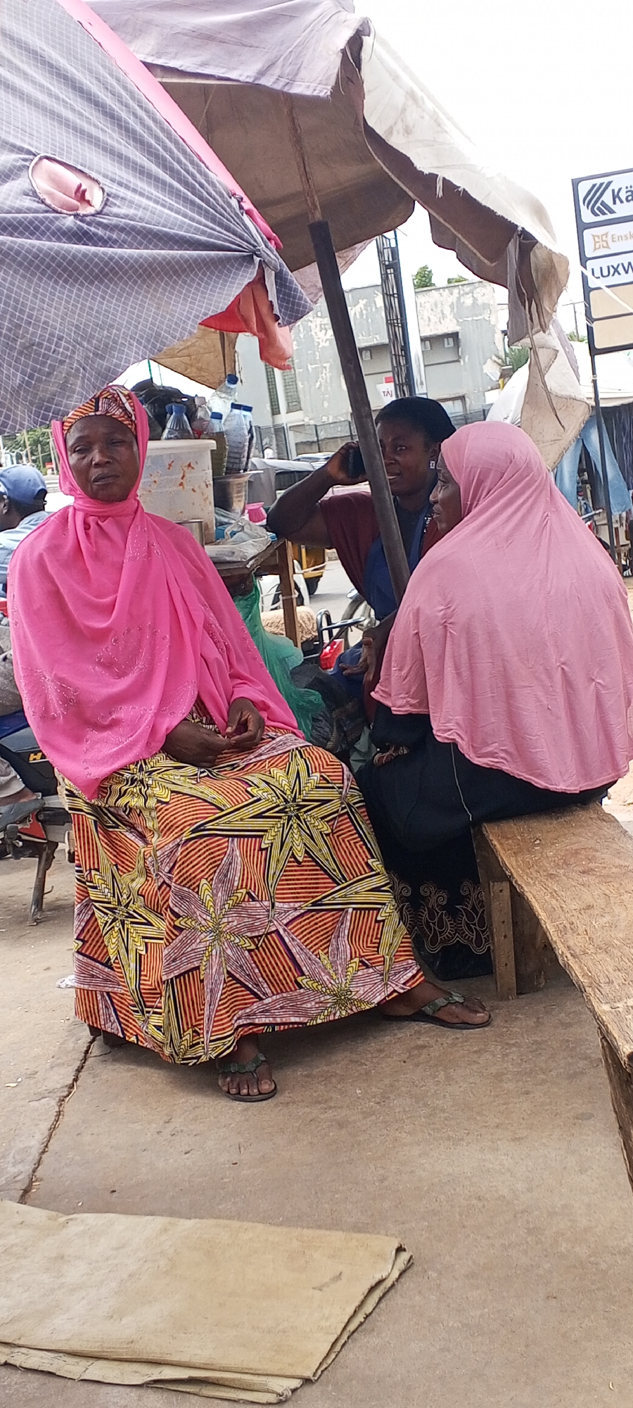 Three women sit outside under a makeshift canopy in Maiburiji, Nigeria. Two of the women are seated on a wooden bench. One is wearing a pink hijab and a long, patterned skirt. The other woman beside her is wearing a pink hijab as well and a dark skirt. A third woman is partially visible behind them, talking on a cell phone. A sign for a business is visible in the background. There is an assortment of items, including bottled water and food containers, sitting on a nearby table. A sheet of rough material lies on the ground in the foreground.