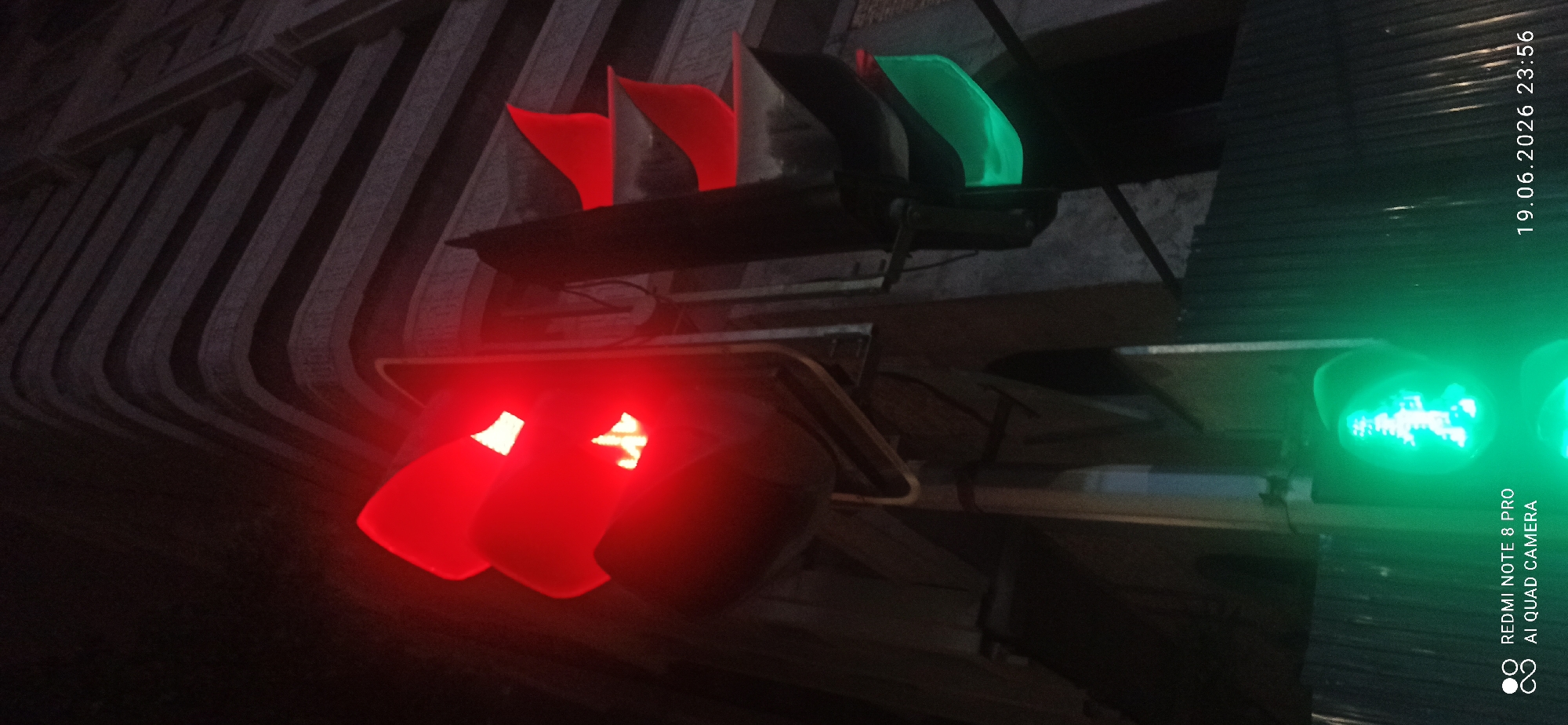 A traffic signal and pedestrian signal are visible at night in an urban environment. The traffic signal has a red light illuminated at the top, a yellow light illuminated in the middle, and a green light illuminated at the bottom. The pedestrian signal shows a green walking figure, indicating it is safe to cross. The background includes a building with a curved facade and horizontal architectural elements. The image also contains timestamps and camera information in the lower left and right corners.