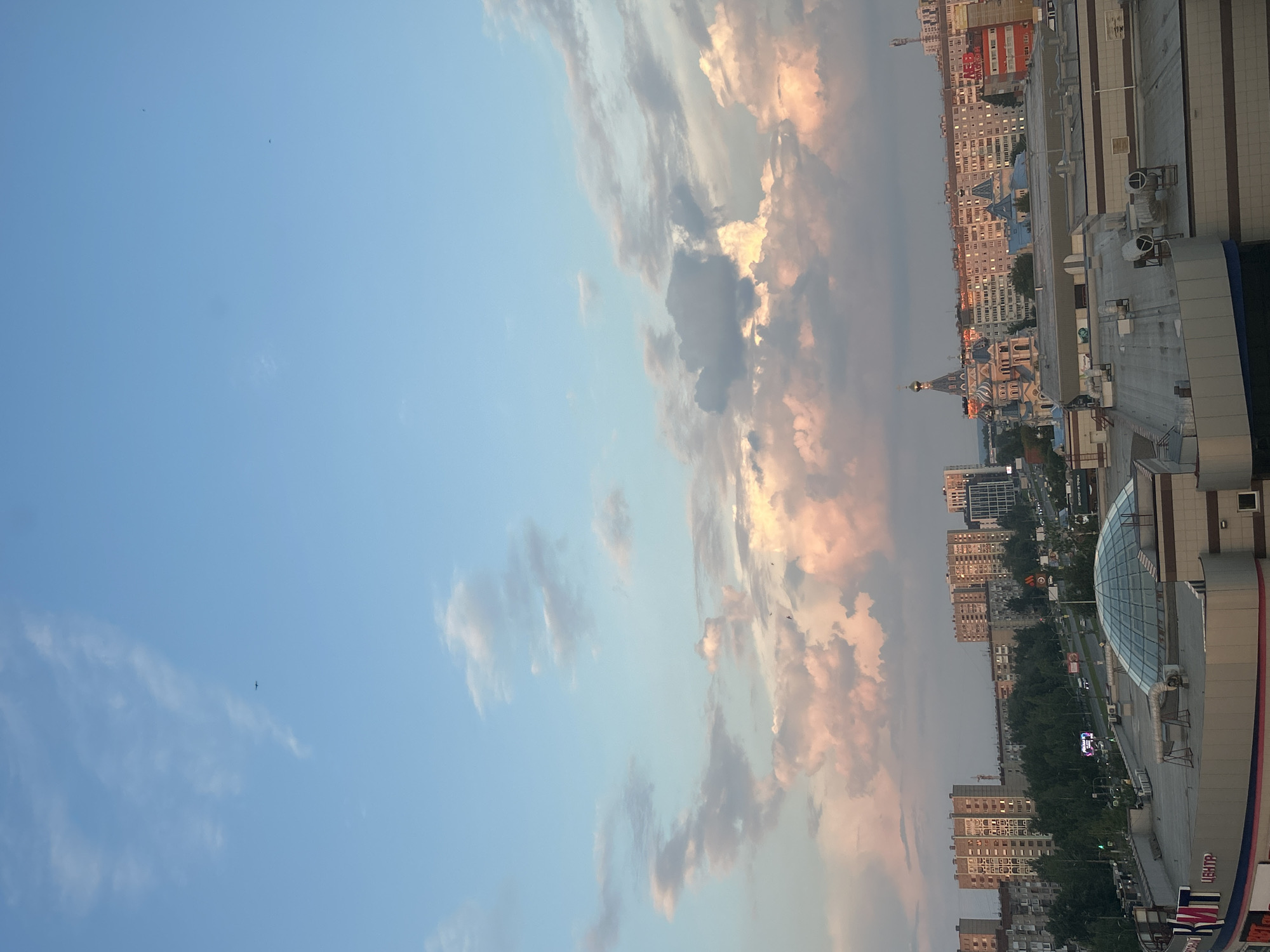 This is an aerial view captured from a high vantage point in Izhevsk, Russia, looking out over a cityscape at dusk. The sky is a gradient of light blue at the top, transitioning to hues of pink and orange from the setting sun illuminating the scattered clouds. In the distance, several apartment buildings and other urban structures are visible, bathed in the warm, soft light. A prominent blue-domed building with intricate architectural details, possibly a church, stands out against the residential buildings. In the foreground, a large, modern building complex with a glass dome is partially visible, suggesting a shopping center or commercial area. The overall scene depicts a tranquil urban environment under a colorful twilight sky.