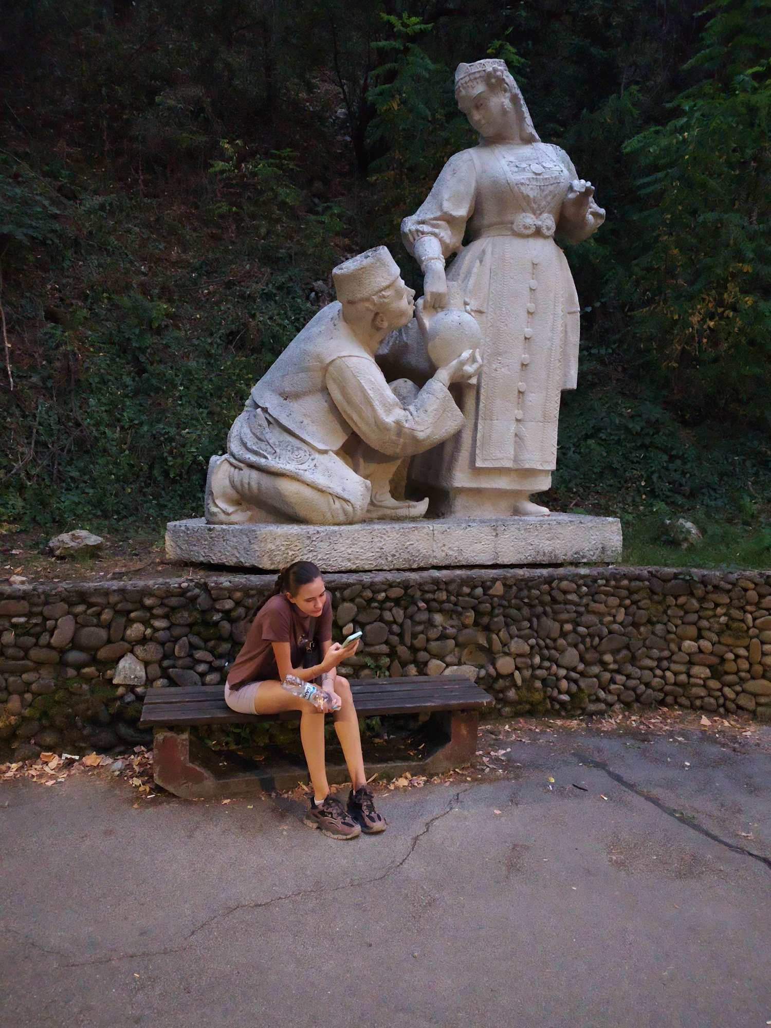 A young woman with brown hair, wearing a maroon t-shirt and light brown shorts, sits on a wooden bench facing right. She is holding a smartphone and a plastic bottle of water. Her feet are adorned with hiking shoes. In the background, a stone wall supports a monument of two stone figures. The male figure kneels, offering a jug to the standing female figure. The monument is surrounded by greenery, suggesting an outdoor setting. The scene appears to be in the city of Lovech, Bulgaria, specifically near the foot of the monument dedicated to the mother of Vasil Levski.