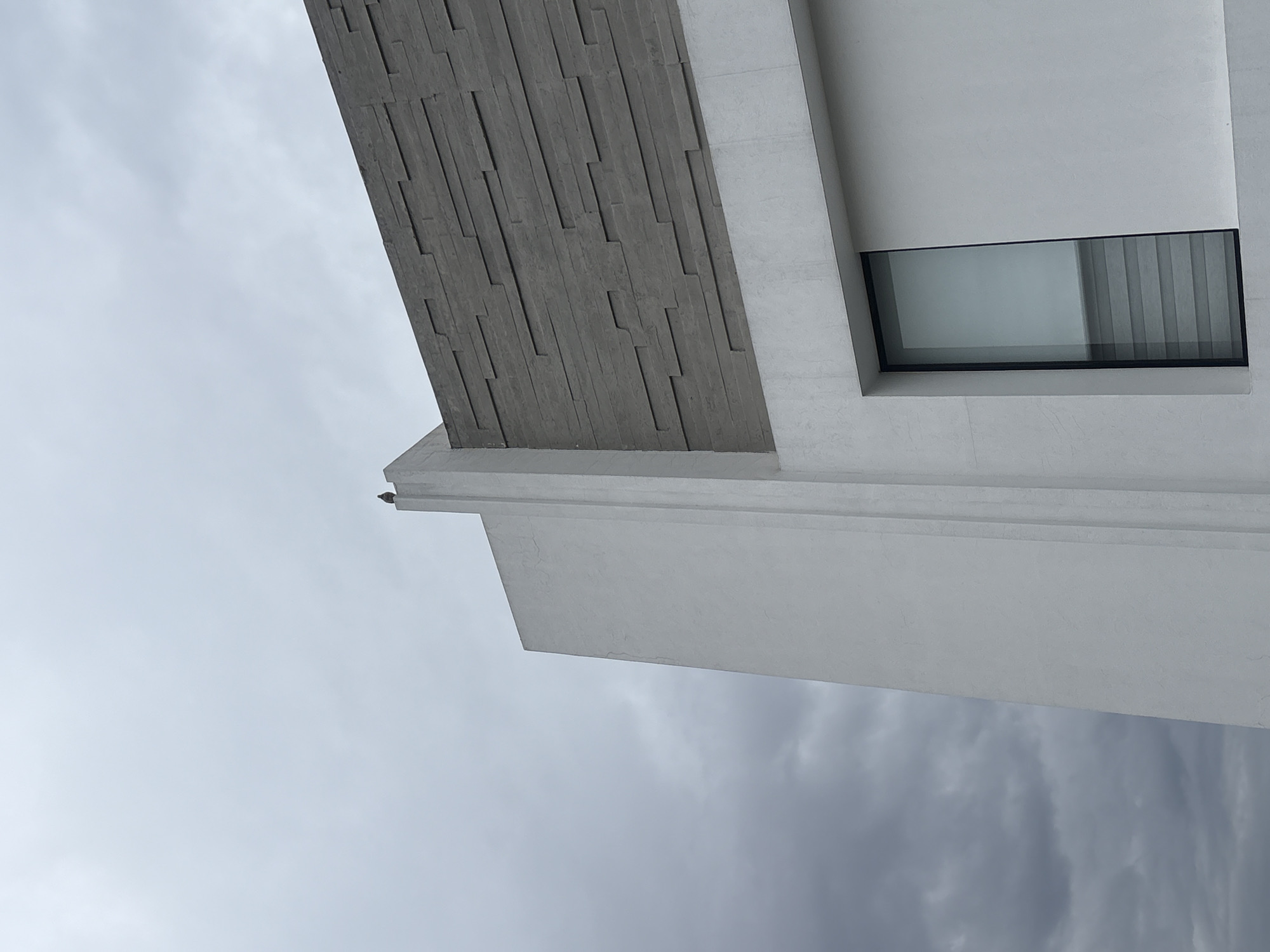 An angled, low-angle shot depicts a section of a modern building against a cloudy sky, possibly in San Bernardino Tlaxcalancingo, Mexico. The building features white walls, a rectangular window, and a section covered in gray, textured stone. The sky occupies the majority of the frame, with the architectural elements filling the upper portion.