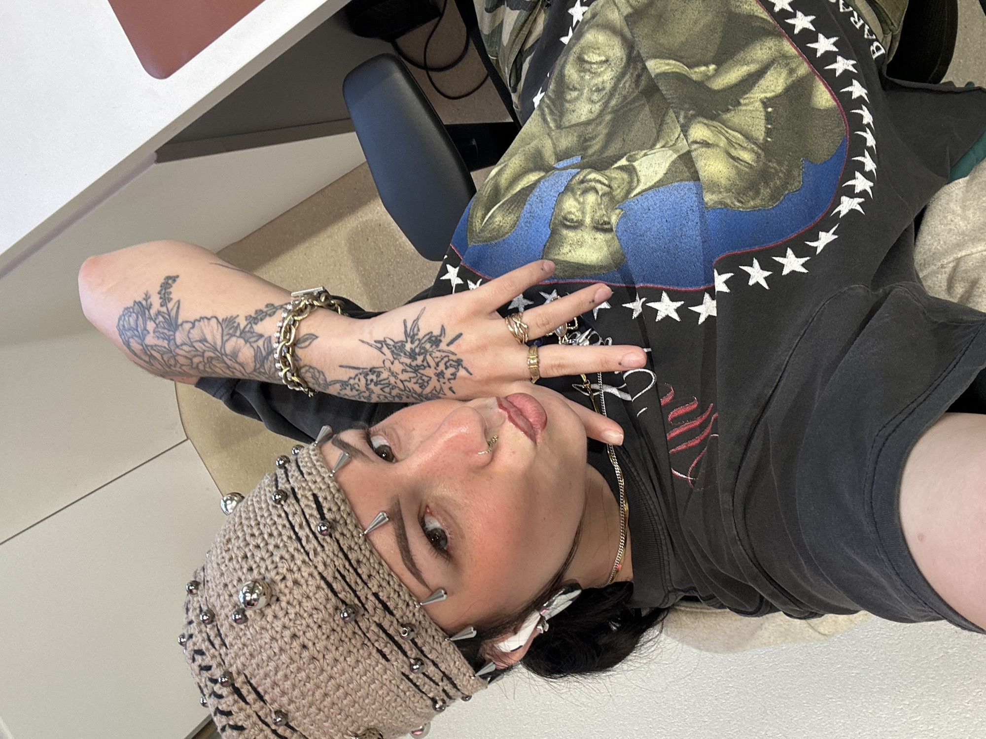 A person wearing a studded crocheted hat is captured in a medium shot. The individual has facial piercings, visible tattoos on their arm, and multiple rings on their fingers. They are wearing a black t-shirt with a graphic design. The background includes a desk and an office chair. The setting does not appear to be in Paris, France.