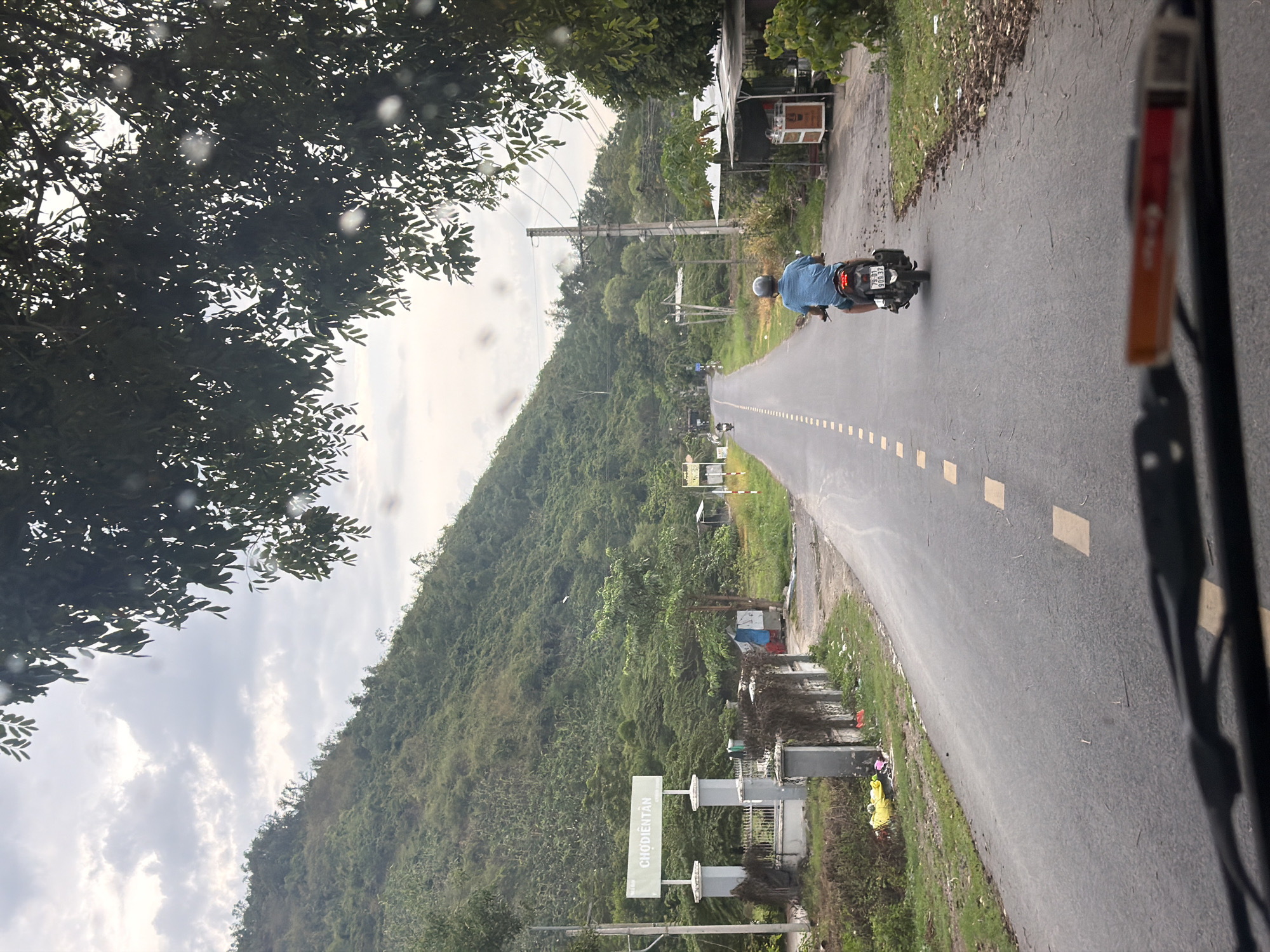 The image captures a road scene in Diên Thọ, Vietnam. A lone motorcyclist, wearing a blue shirt and helmet, rides away from the camera on a paved road. The road is marked with dashed yellow lines and bordered by a grassy verge. On the left side of the road, a sign with the text "CHỢ DIỆN TÂN" is visible, supported by two grey pillars. Beyond the sign, a white fence and some vegetation can be seen. The background features a lush, green, mountainous landscape covered in trees. On the right side of the road, there is a small structure with a white roof, appearing to be a roadside stall. The sky is overcast with clouds. The perspective suggests the photo is taken from a vehicle.