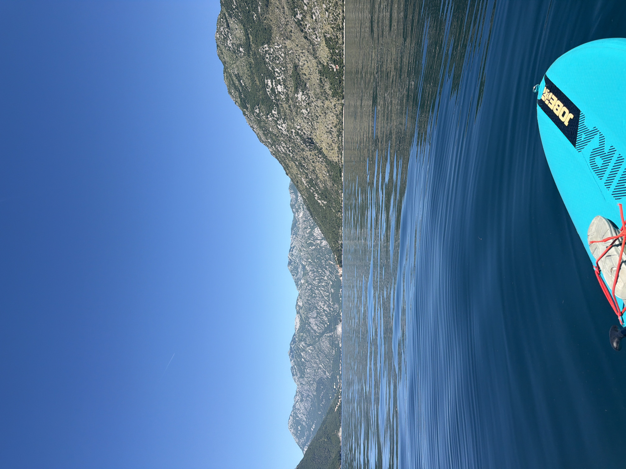 The image shows a section of the Bay of Kotor in Stoliv, Montenegro, taken from a low angle at water level. The sky is a clear, unblemished blue. A portion of mountainous terrain rises on the left, composed of rock and patchy vegetation. Water ripples cover the right half of the image. On the far right is a portion of a turquoise stand-up paddleboard with a "JOBE" logo.