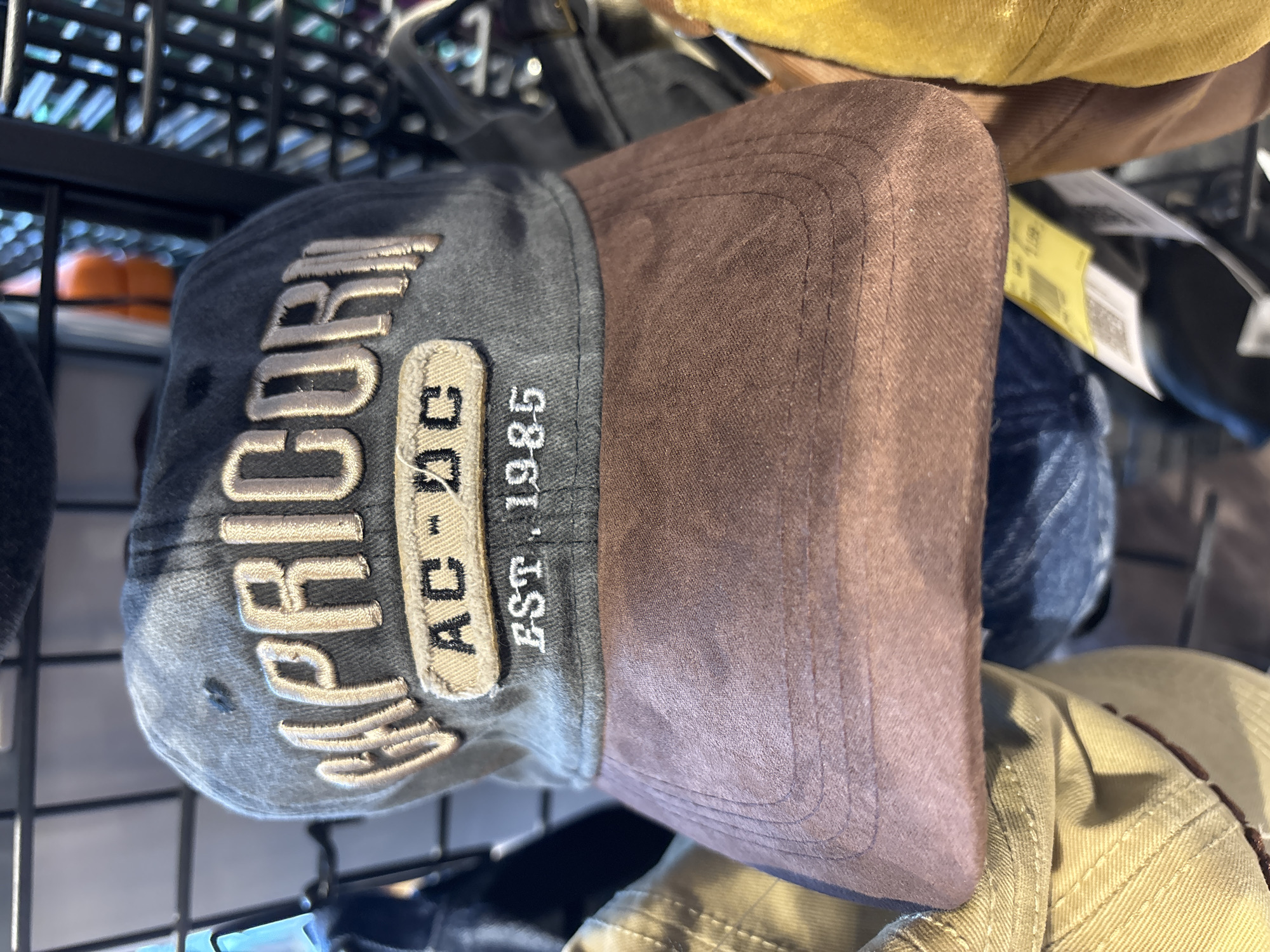 A close-up shot shows a brown and grey baseball cap with "CAPRICORN" embroidered in gold lettering across the front. Below "CAPRICORN" are two smaller embroidered patches, the top one reads "AC-DC" and the bottom one "EST. 1985". The cap is hanging on a black metal display rack, likely in a retail store. The background is blurred, but suggests other items of clothing or merchandise. There is no indication of the city of Stimul, Russia.