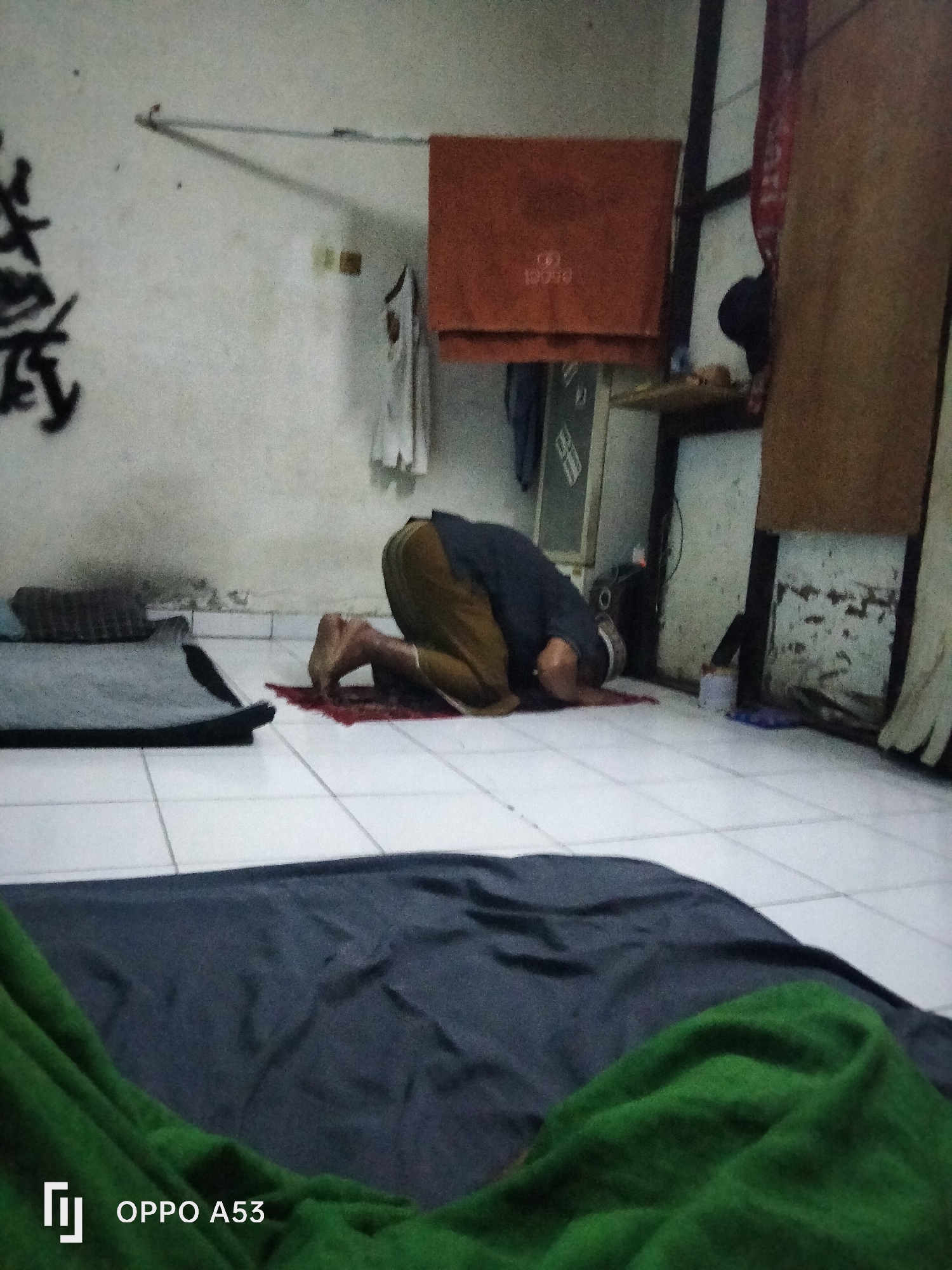 A low-angle, dimly lit, full-shot photograph captures a person performing the Islamic prayer known as *sujud* (prostration) on a red prayer mat. The individual is seen from the knees down, with their forehead touching the mat. They are wearing tan pants and a dark shirt. The room appears to be sparsely furnished with a white tiled floor. To the left of the prayer mat, a grey mat is visible, along with a dark grey blanket and a green blanket in the foreground. The background reveals a roughly textured white wall with some dark graffiti on the far left. A small orange object, possibly a towel or a cloth, hangs on the wall above the person. Further to the right, a wooden shelf holds a few indistinct items. The overall impression is of a simple, humble living space. The image is tagged with "OPPO A53," indicating it was taken with a mobile phone. The location is identified as Denpasar, Indonesia.
