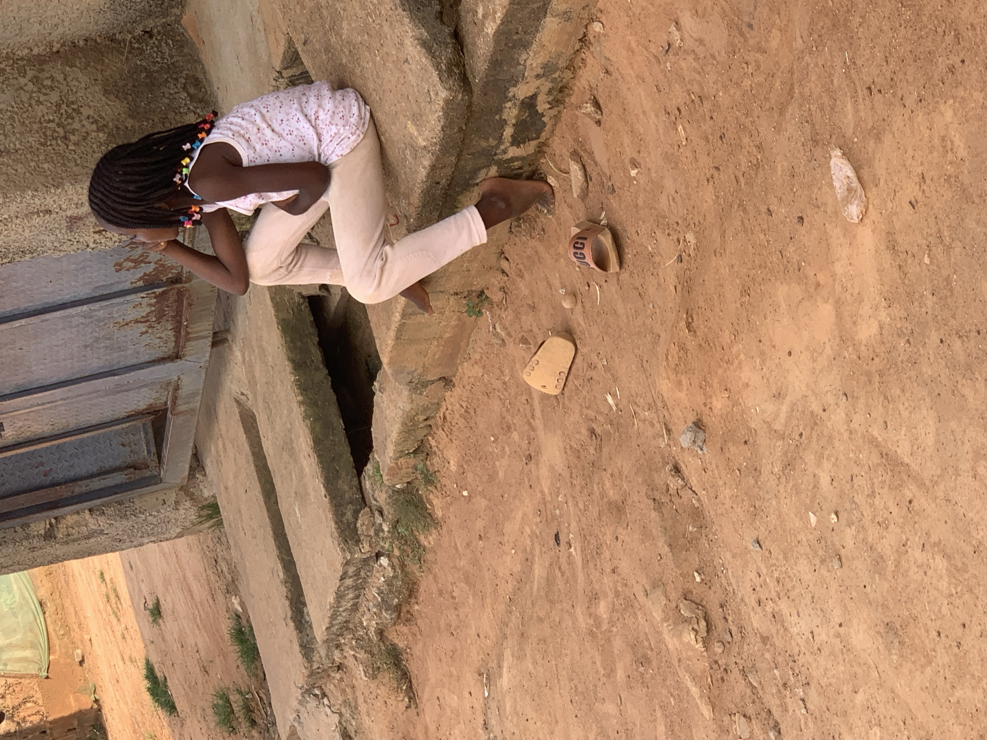 A young girl with braided hair adorned with colorful beads sits on a concrete ledge outdoors. She is wearing a white sleeveless top with a pink floral pattern and light-colored pants. Her right hand supports her head as she looks to her right, appearing contemplative. Nearby, on the sandy ground, are two slides, one tan and one brown with "GUCCI" visible on it. The environment appears to be a dusty, outdoor area with rough concrete structures and sparse vegetation. The scene is likely in Sabon Tasha, Nigeria, given the context.