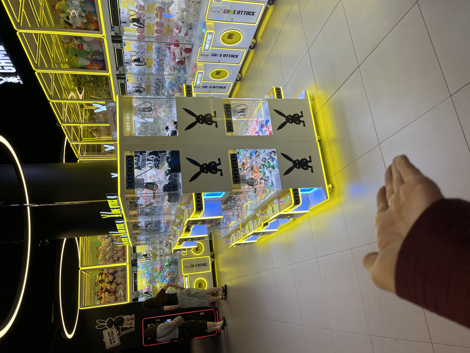 The image shows the interior of a game arcade, likely a "Game Attack" arcade based on the signage on the machines. The arcade features multiple rows of prize claw machines, filled with plush toys and small figurines. The machines are brightly lit with yellow and blue neon lights, creating a vibrant atmosphere. Two individuals, a woman wearing a grey top and patterned skirt and another woman in a black top and light-colored shorts, are visible standing in the foreground, observing the machines. The floor is tiled with a light-colored pattern. In the upper portion of the image, more claw machines are visible, filled with an assortment of plush toys, including rabbits and other cartoon characters. A sign with a cartoon rabbit and Russian text, "ОБМЕН ДЕНЕГ НА ЖЕТОНЫ" (exchange money for tokens), is mounted on the wall. The lighting suggests an indoor environment, possibly at night. The presence of "GAME ATTACK" branding and the style of the arcade machines are consistent with venues found in tourist areas. While the specific location in Sochi, Russia is not definitively identifiable from the visual cues alone, the type of establishment and the Russian signage suggest it is likely within Russia, and potentially in a city like Sochi where such entertainment venues are common.
