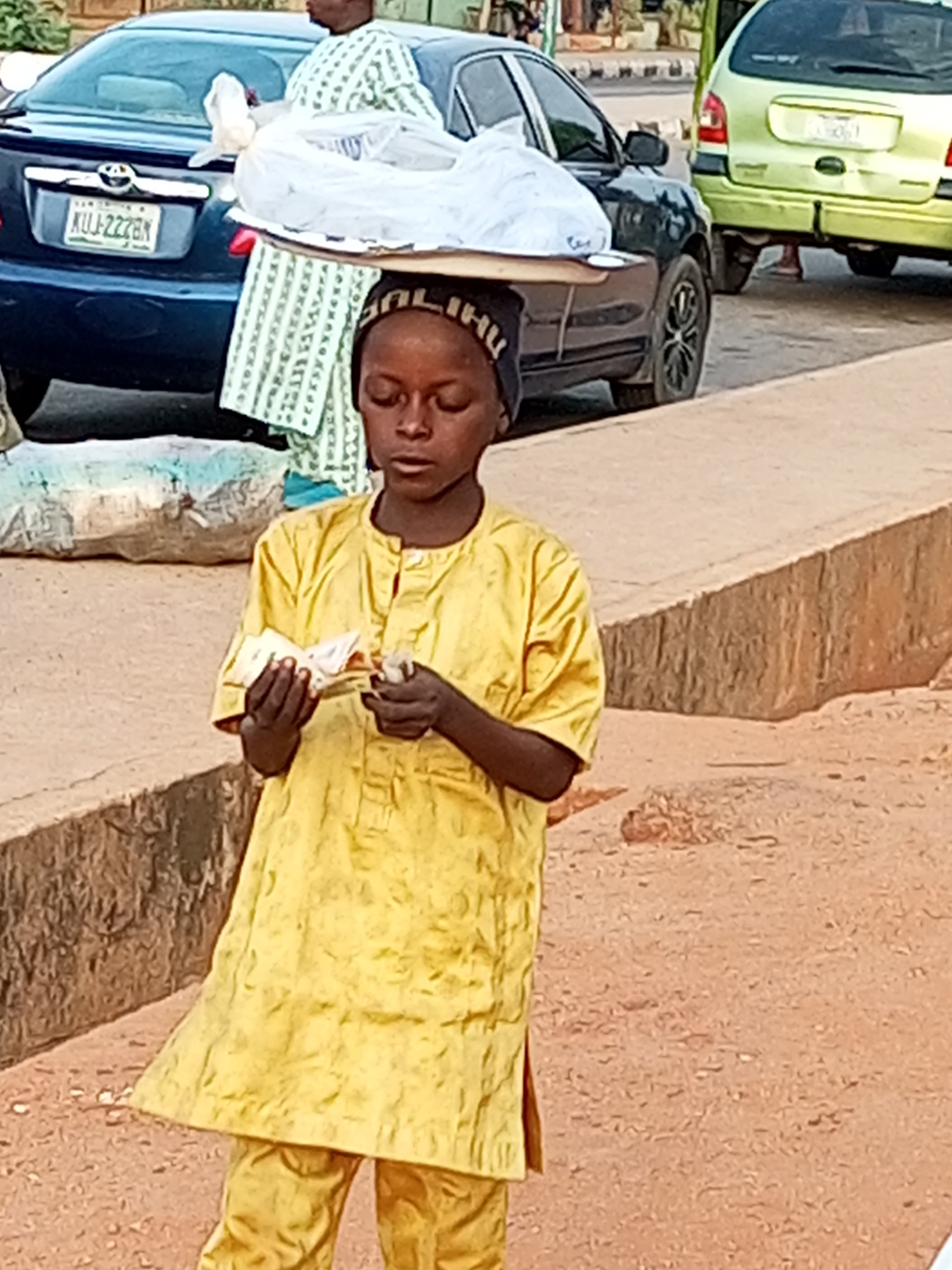 A young boy, likely of Nigerian descent, stands in the foreground wearing a bright yellow traditional outfit, including a tunic and pants. He is holding a wad of cash in his hands, appearing to be counting or examining it. On his head, he balances a tray piled high with white plastic-wrapped goods. The boy is positioned in front of a dark blue sedan with a license plate reading "KUJ-2228N." Behind the sedan is a pale green hatchback, and further in the background, buildings and other vehicles are visible. The scene takes place on a dirt or gravel surface, suggesting an outdoor setting, possibly a street or market area in Gombe, Nigeria. The lighting indicates it is daytime.