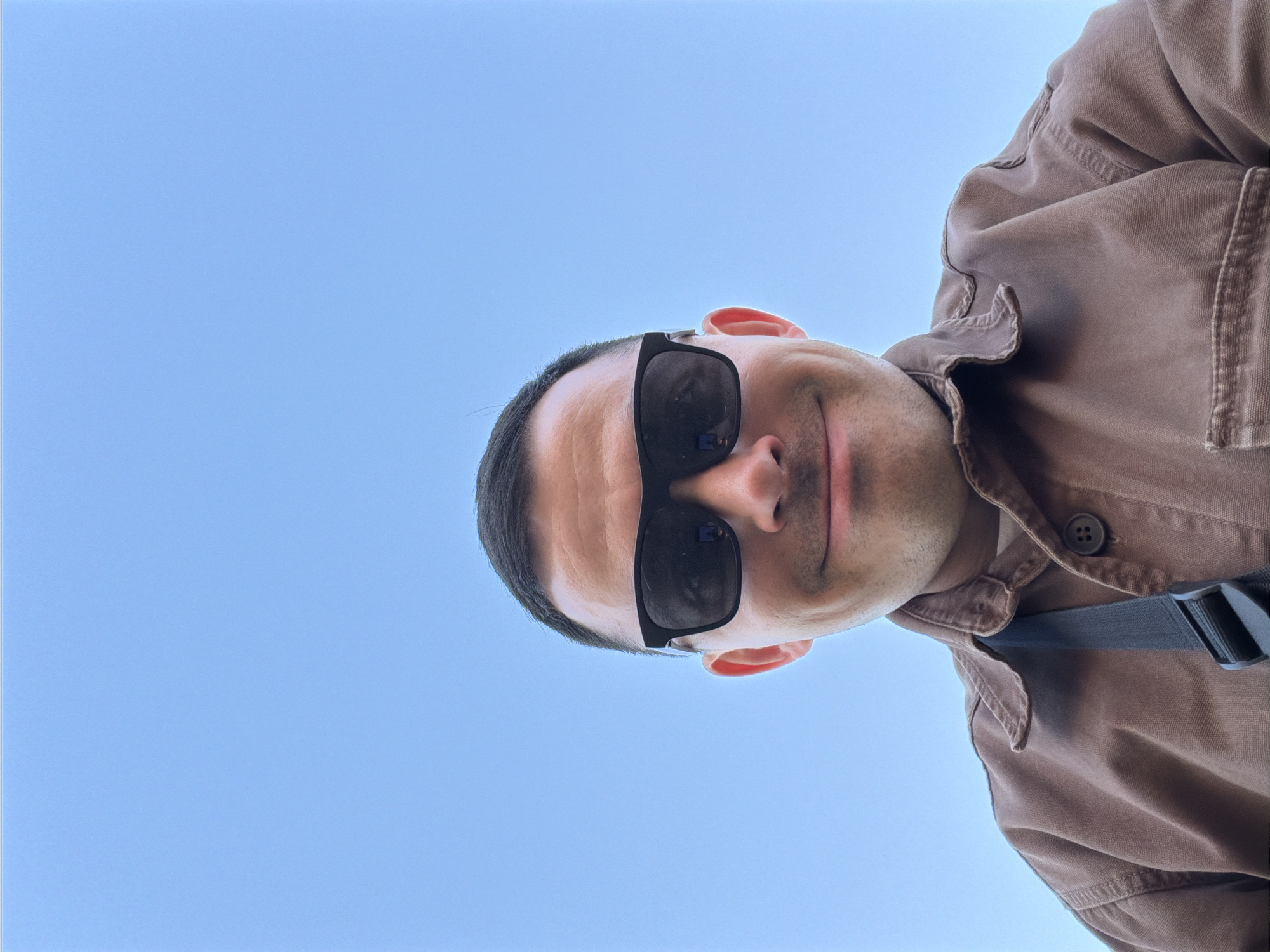 A low-angle shot of a man with short dark hair and sunglasses looking down at the camera. He is wearing a brown shirt or jacket, and a black strap is visible across his chest. The background is a clear blue sky. There is insufficient information to determine the location as Izhevsk, Russia.