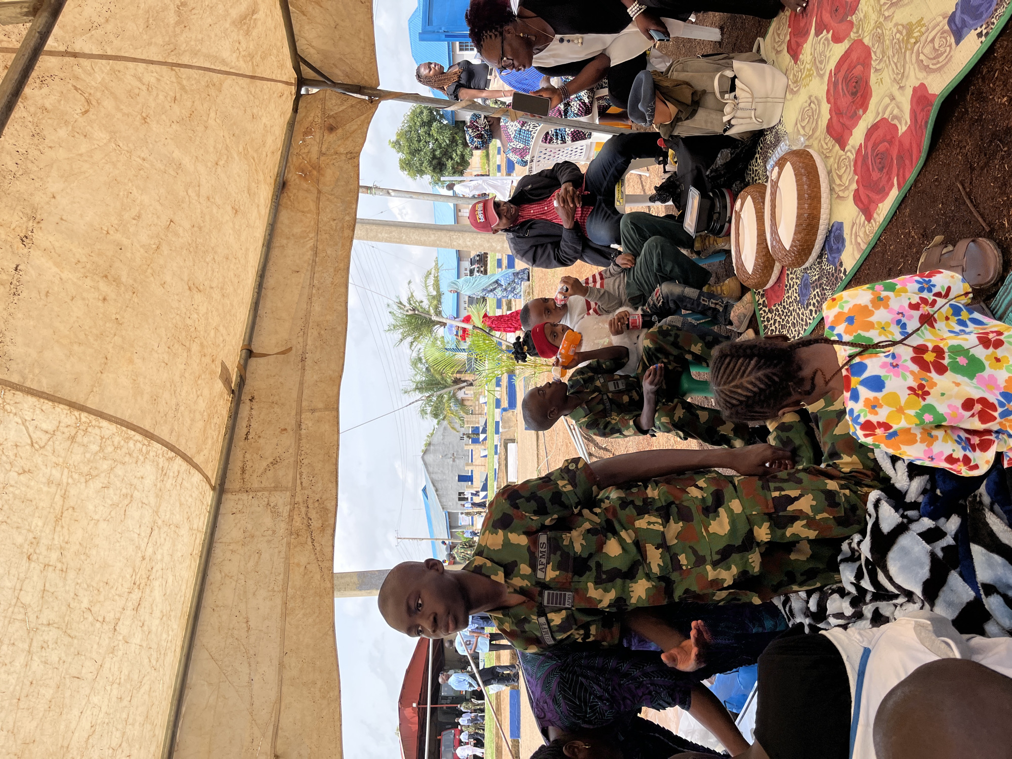 This media file captures an outdoor gathering under a temporary shelter, likely a tent, with a rough, tan-colored tarp overhead. The scene appears to be in Bar Jirgi Summa, Nigeria. The ground is covered with dirt and debris.
Several people are present, some seated on the ground on colorful mats, others on chairs. Many are wearing camouflage military uniforms, suggesting a military event or gathering. One individual in the foreground wears a camouflage uniform with "AFMS" visible on a patch. Another person in the mid-ground is wearing a red and black plaid shirt and a red baseball cap. The people are a mix of ages and genders.
Objects visible include various bags, some appearing to be backpacks and purses, as well as two round, cushion-like objects with a brown and white pattern. There are also water bottles and a drink in a plastic bottle. The overall atmosphere seems relaxed and informal, with people interacting and some looking towards the camera.
The lighting suggests it is daytime, with the sun casting shadows. The sky is visible in some parts of the image and appears to be overcast or hazy. In the background, there are buildings with blue accents, possibly a school or institutional facility, and some trees. The presence of military personnel and the informal gathering under a shelter indicate a community event or a break in a military activity.