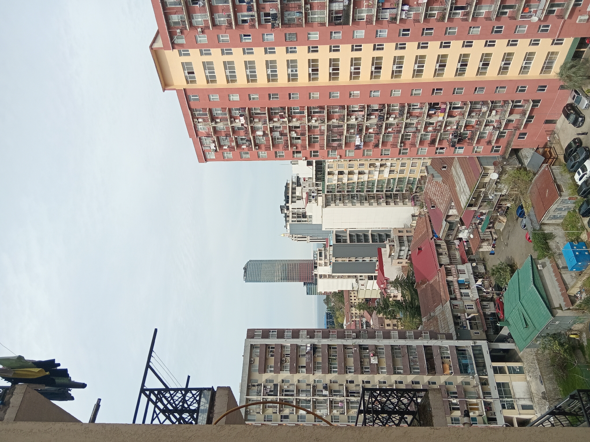 The image shows an urban scene in Batumi, Georgia. A high-angle view from a rooftop captures a panorama of buildings, including several high-rise apartment complexes with numerous windows and balconies. One prominent building is peach and red, while another is brown and tan. In the background is a tall skyscraper. Below the high-rises are older, smaller buildings with red and green roofs. Some cars are visible in the street, and various satellite dishes and antennas are seen on the buildings. The sky is overcast. Laundry hangs from some balconies.