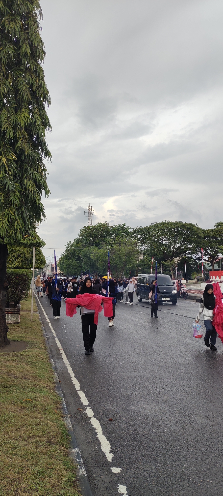 This is a vertical, eye-level, medium shot of a parade on a street in Lhokseumawe, Indonesia, likely on Jalan Merdeka. The sky is overcast and gray, and the asphalt road is wet.
In the foreground, a young woman wearing a black hijab and black clothing walks forward, holding a large, folded piece of red fabric. To her right, another young woman wearing a black hijab and a striped shirt carries several plastic bottles and another piece of red fabric.
Behind them, a procession of people, mostly young women wearing hijabs and dark clothing, walk down the street. Some carry tall, slender flags with colored tops, while others hold red fabric pieces. A dark blue van is visible in the middle ground, driving in the same direction as the parade. A motorcycle is also visible further down the road.
On the left side of the image, a tall tree with dense green foliage lines a grassy area with a concrete curb. In the background, there are more trees and a tall cellular tower. The overall impression is of a community event taking place under a cloudy sky.