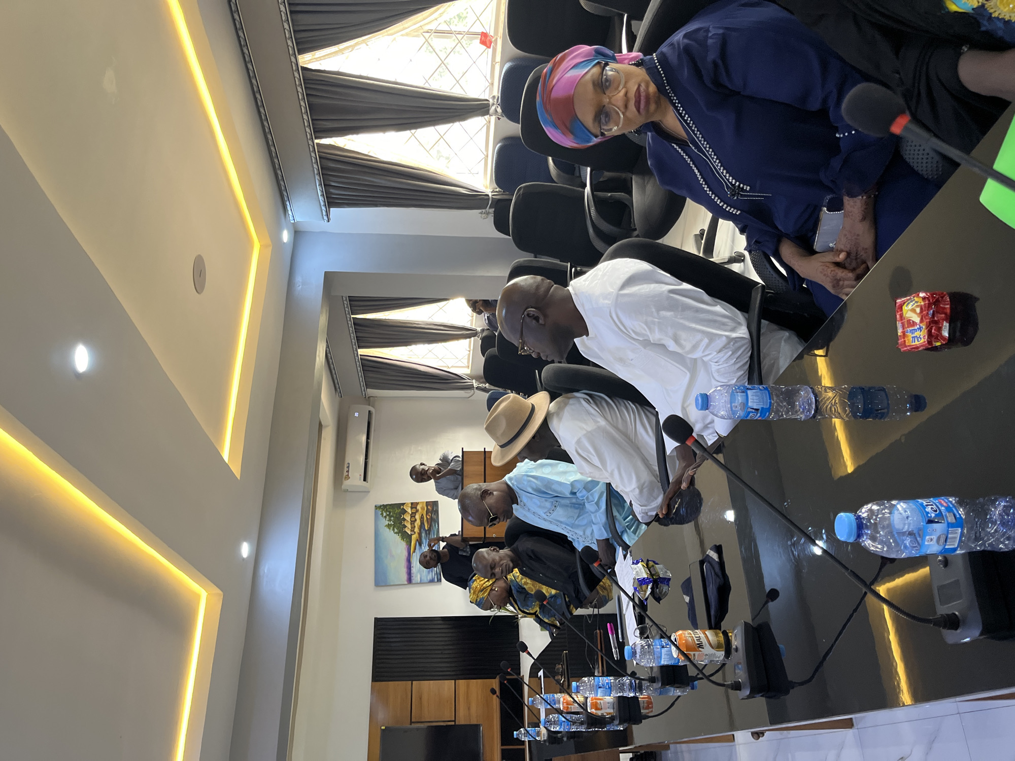 This media file depicts a meeting taking place in what appears to be a conference room in Jos, Nigeria. The scene is dominated by a long, dark, reflective table around which several individuals are seated.
The subjects are primarily people, diverse in age and gender, dressed in formal and traditional attire. A woman in a blue outfit and headscarf, wearing glasses, is prominent in the foreground. Other individuals are seen wearing white, blue, and patterned clothing, some with hats. Microphones are placed in front of many of the seated individuals, suggesting an organized discussion or presentation.
On the table, several water bottles are visible, along with a packet of what appears to be snacks or candy, and electronic devices like a speaker. The room itself features modern lighting with yellow LED strips illuminating the ceiling. Dark curtains cover windows in the background, and an air conditioning unit is visible on the wall, along with a painting.
The activity appears to be a formal meeting or conference. The individuals are mostly looking down or towards each other, engaged in the proceedings. The lighting suggests it could be daytime, but the indoor setting makes it difficult to ascertain the exact time of day or weather conditions. There are no explicit emotional cues visible, but the overall atmosphere seems serious and focused.
Visible text includes labels on the water bottles and the packaging of the snacks, but they are not clearly legible. The overall impression is of a professional gathering in an indoor, well-equipped meeting space.