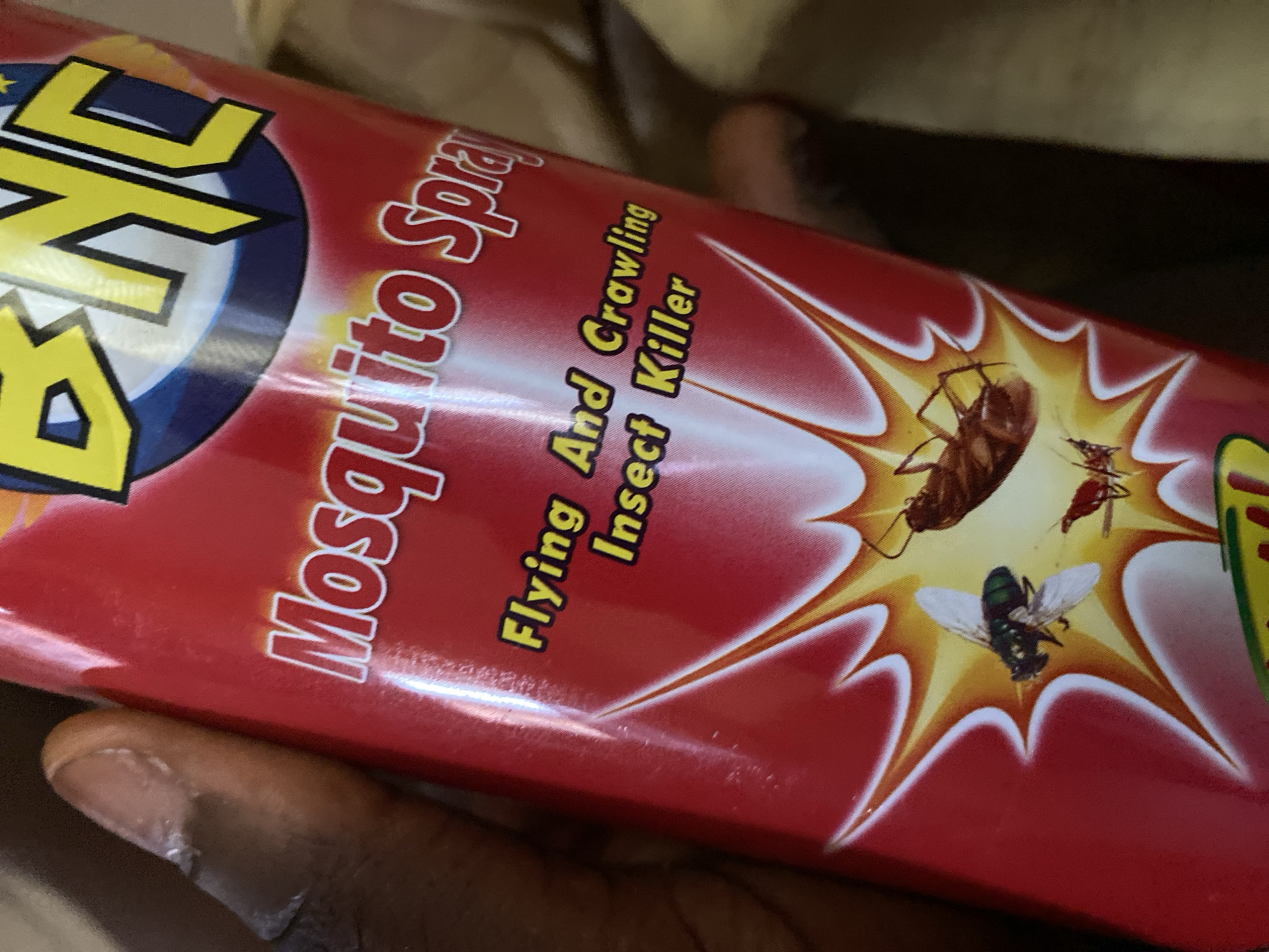 The image shows a close-up, angled shot of a red can of "NYC Mosquito Spray." The can has yellow and blue lettering and graphics. Text on the can reads, "Mosquito Spray," "Flying And Crawling Insect Killer," and "NYC." The spray depicts stylized images of insects, including a cockroach, a fly, and what appears to be a mosquito or gnat. The background is out of focus, showing muted earth tones. A person's thumb and fingers are visible in the lower left corner, holding the can. The lighting suggests an indoor setting with artificial light. The text indicates the product is an insecticide for flying and crawling insects. The provided location context is Jauro Yinu, Nigeria.