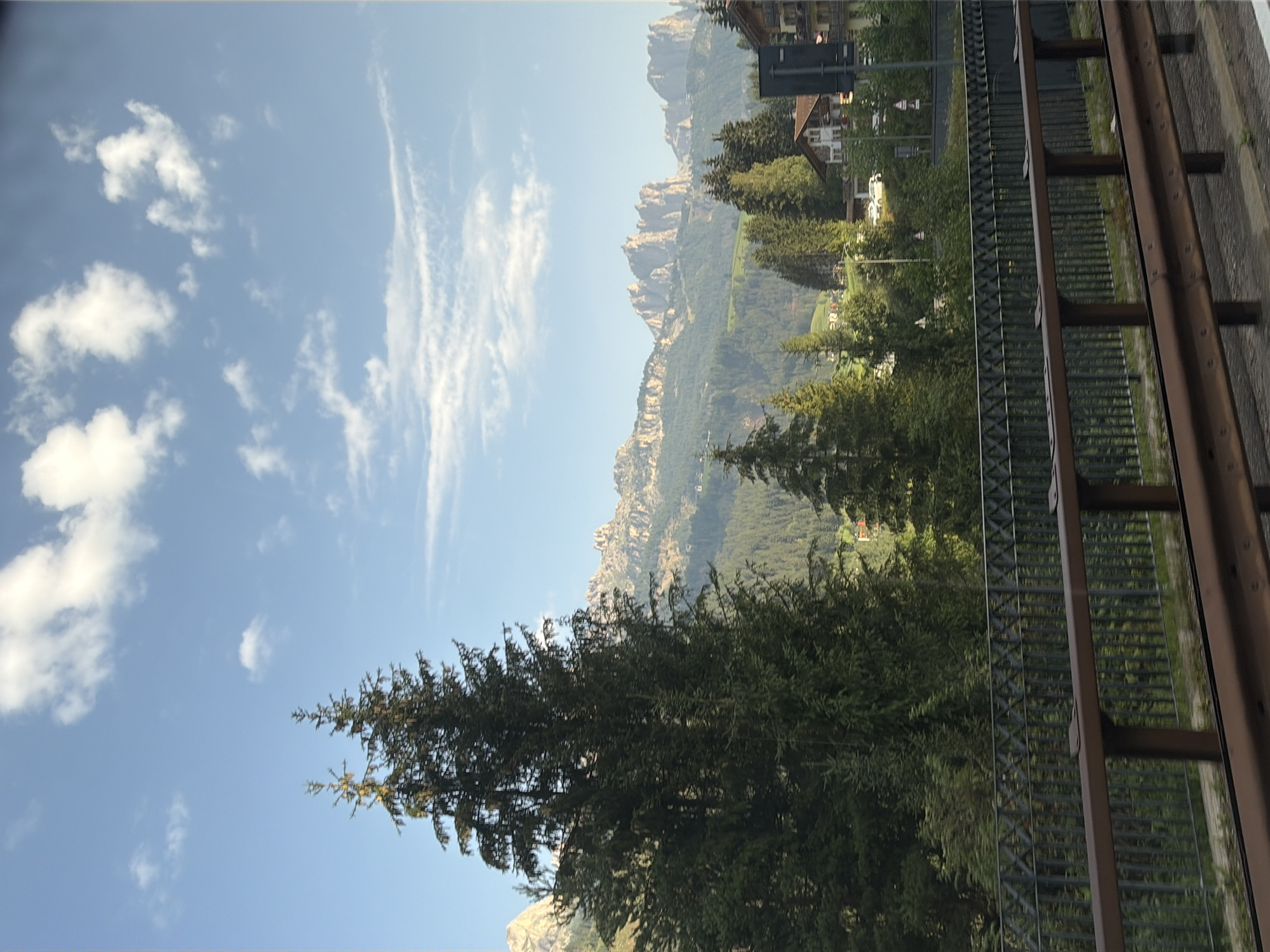 The image shows a scenic view of Soraga di Fassa, Italy, on a clear day with scattered clouds. In the foreground, a metal railing with a brown top beam separates the viewer from a dense green forest. The railing has a pattern of interwoven metal bars. Beyond the railing, tall evergreen trees dominate the landscape, their dark green foliage contrasting with the lighter green of distant hills.
In the mid-ground, the village of Soraga di Fassa is visible nestled amongst the trees. Traditional alpine houses with pitched roofs are scattered throughout the area. A white car is parked on a road in front of one of the houses. Further in the background, majestic mountain peaks with rugged, rocky formations rise sharply against the blue sky. The mountains are a light, sandy color and appear to be lit by the sun, highlighting their textured surfaces. The overall impression is one of natural beauty and a peaceful mountain village setting.