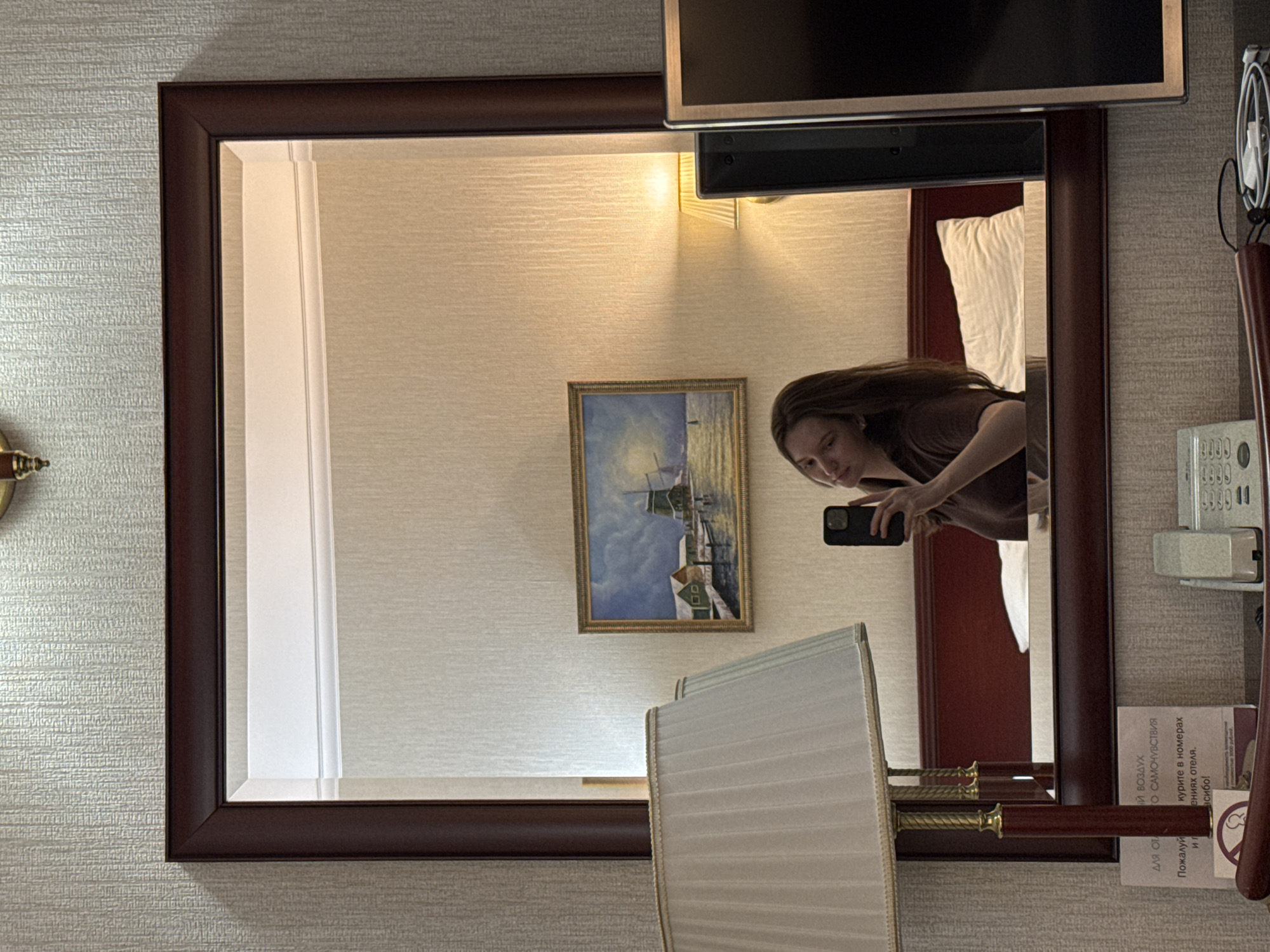The image appears to be a reflection in a mirror within a hotel room. A young woman is visible in the mirror, taking a selfie with her phone. She has long, brown hair and is wearing a brown shirt. Behind her, a painting of a windmill and houses by the water is hung on the wall. A bedside lamp with a pleated shade is also visible. In the foreground, a phone and some charging cables are on a table. The room has textured wallpaper and a dark wooden mirror frame. The provided location, Журавлево (кемеровский), Russia, suggests this scene is set in a hotel room in that specific region.