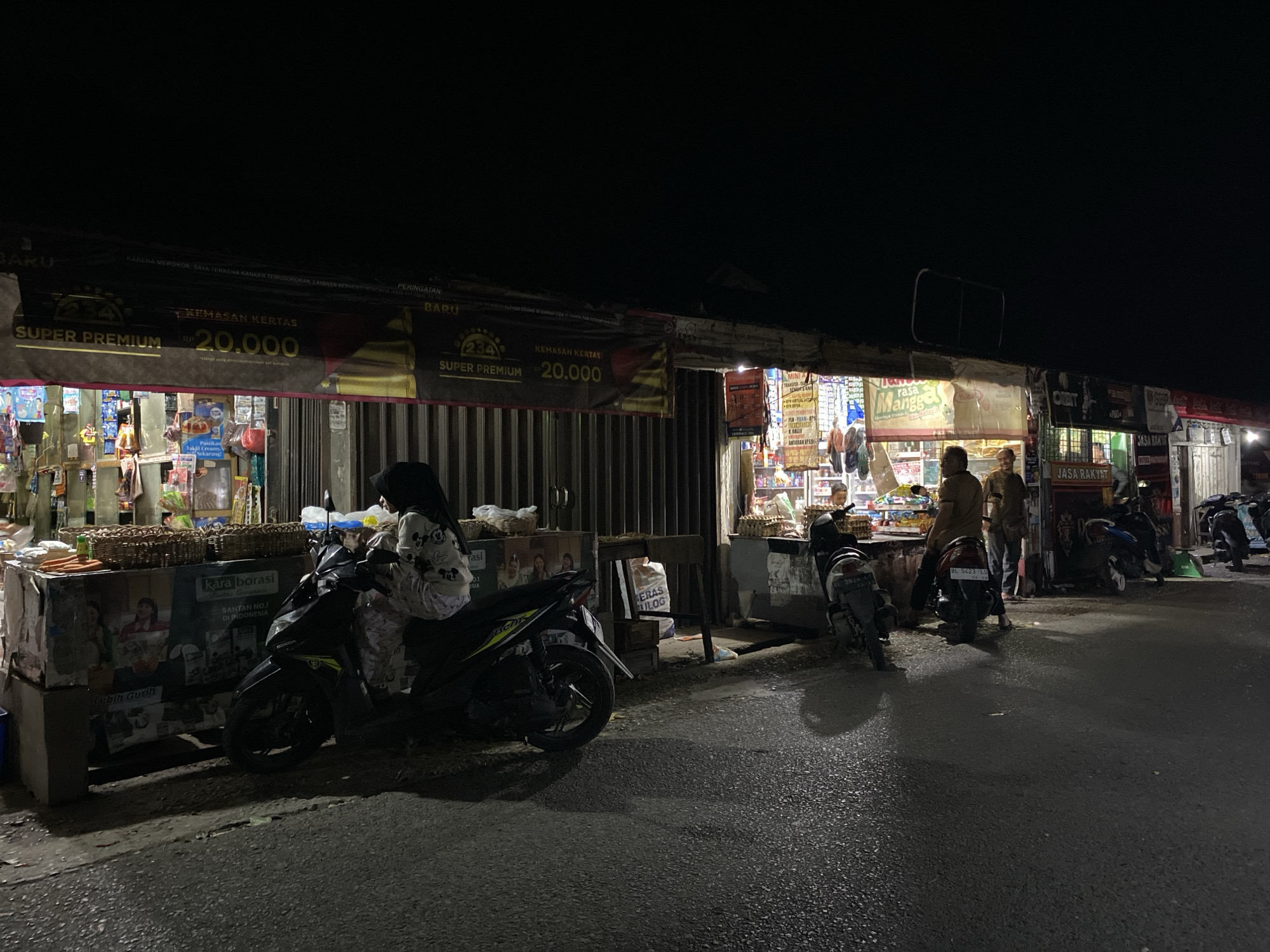 This is an outdoor, nighttime photograph of a street-side market in Aneuk Paya, Indonesia. The scene is illuminated by artificial lights from the market stalls and the headlights of parked motorcycles. In the foreground, the asphalt road reflects the ambient light, with shadows cast by several parked motorcycles.
The market stalls are arranged along the side of the road. The stalls appear to be covered by awnings or tarps, with goods displayed on shelves and tables. One stall prominently features a banner with "SUPER PREMIUM RP 20.000" in large lettering. Various products, including packaged goods and what appear to be woven baskets filled with items, are visible in the stalls.
Several people are present. A woman wearing a hijab and patterned clothing is seated on a black motorcycle on the left side of the image. To her right, a man in a light brown shirt and dark pants stands near a motorcycle. Further down the market, two more men are standing near a stall. Other motorcycles are parked in front of the stalls, and some appear to be in motion or have their lights on. The overall environment suggests a bustling local market scene at night.
