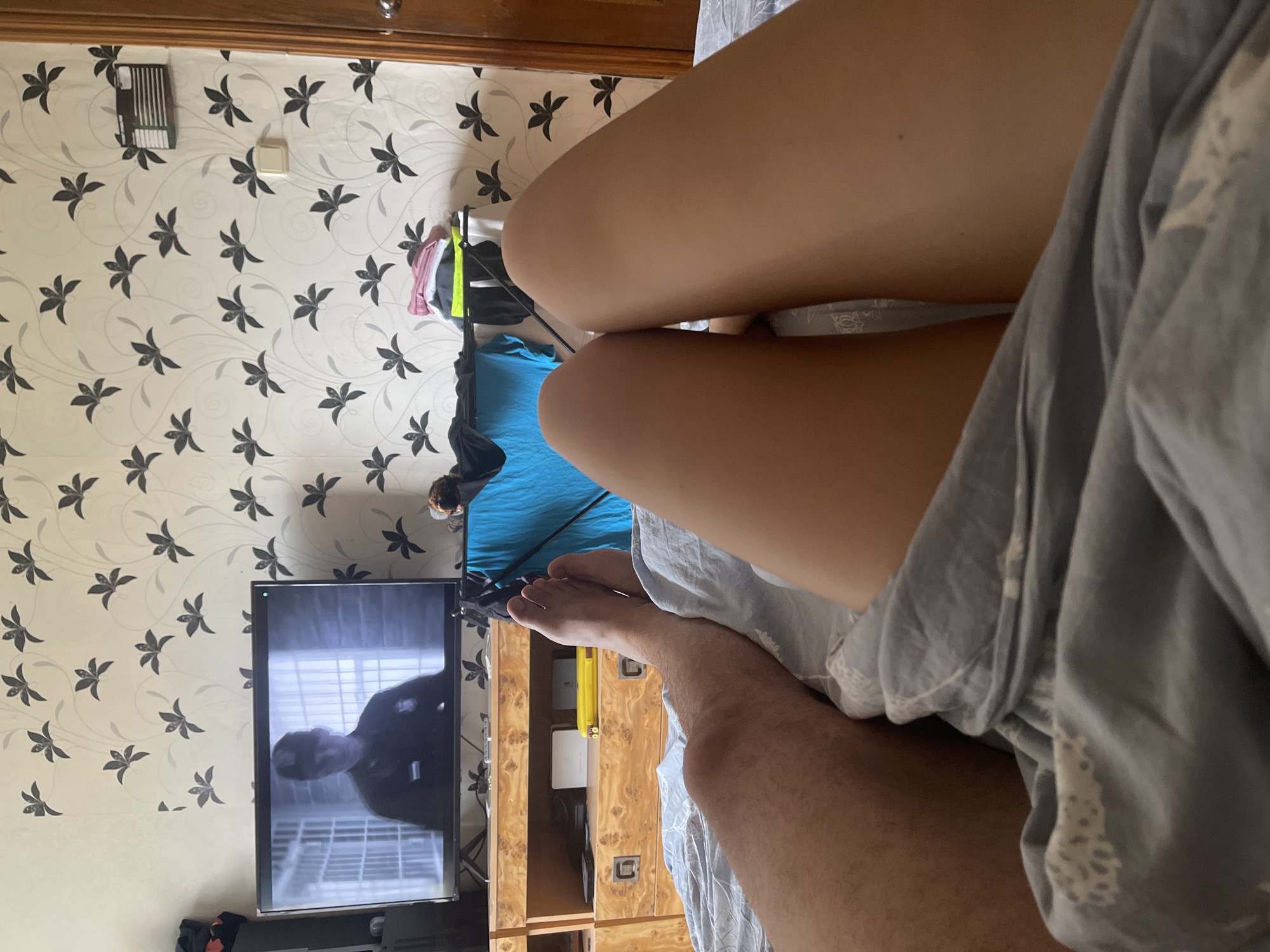 The image shows a bedroom interior with a television set displaying a scene from a movie. The camera is positioned low, looking up at the television and across two individuals' legs lying on a bed. The room is decorated with floral wallpaper. One person's legs, appearing to be male, are visible on the left side of the frame, covered by a grey duvet. The other person's legs, appearing to be female, are on the right side, also covered by the duvet. The television is on a wooden entertainment unit, beneath which are several drawers and some objects. On the wall behind the television, there is a shelf with some folded clothes. The overall scene depicts a relaxed atmosphere, likely a home environment in the city of Shcherbinka, Russia, where the individuals are engaged in watching television.