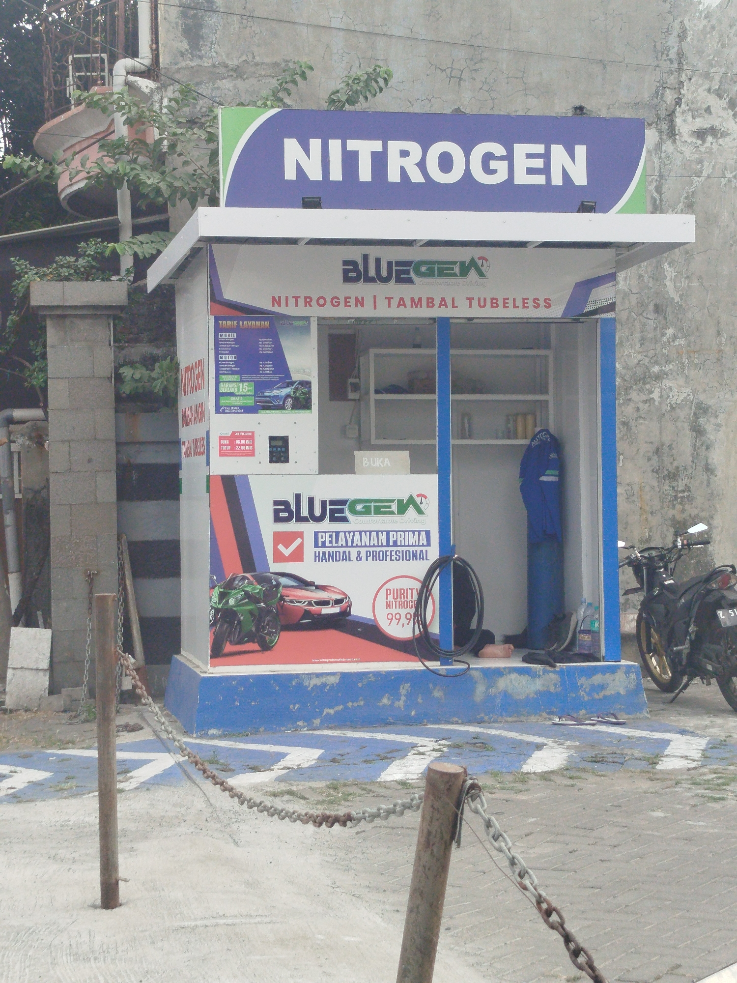 The image depicts a nitrogen filling station in Yosowilangun, Indonesia. A blue and white structure with a large "NITROGEN" sign is the central focus. The station appears to be a small kiosk with an open front, revealing shelves with various items, a blue gas cylinder, and a person in blue overalls lying on the ground. A hose is coiled on the ground in front of the kiosk. A red sports car and a green motorcycle are depicted on promotional material on the side of the kiosk. A black motorcycle is parked to the right of the kiosk. The ground is paved with a pattern of blue and white chevrons, and a rusty chain is draped across the foreground, attached to a metal post. The background shows a weathered concrete wall and some overgrown vegetation.