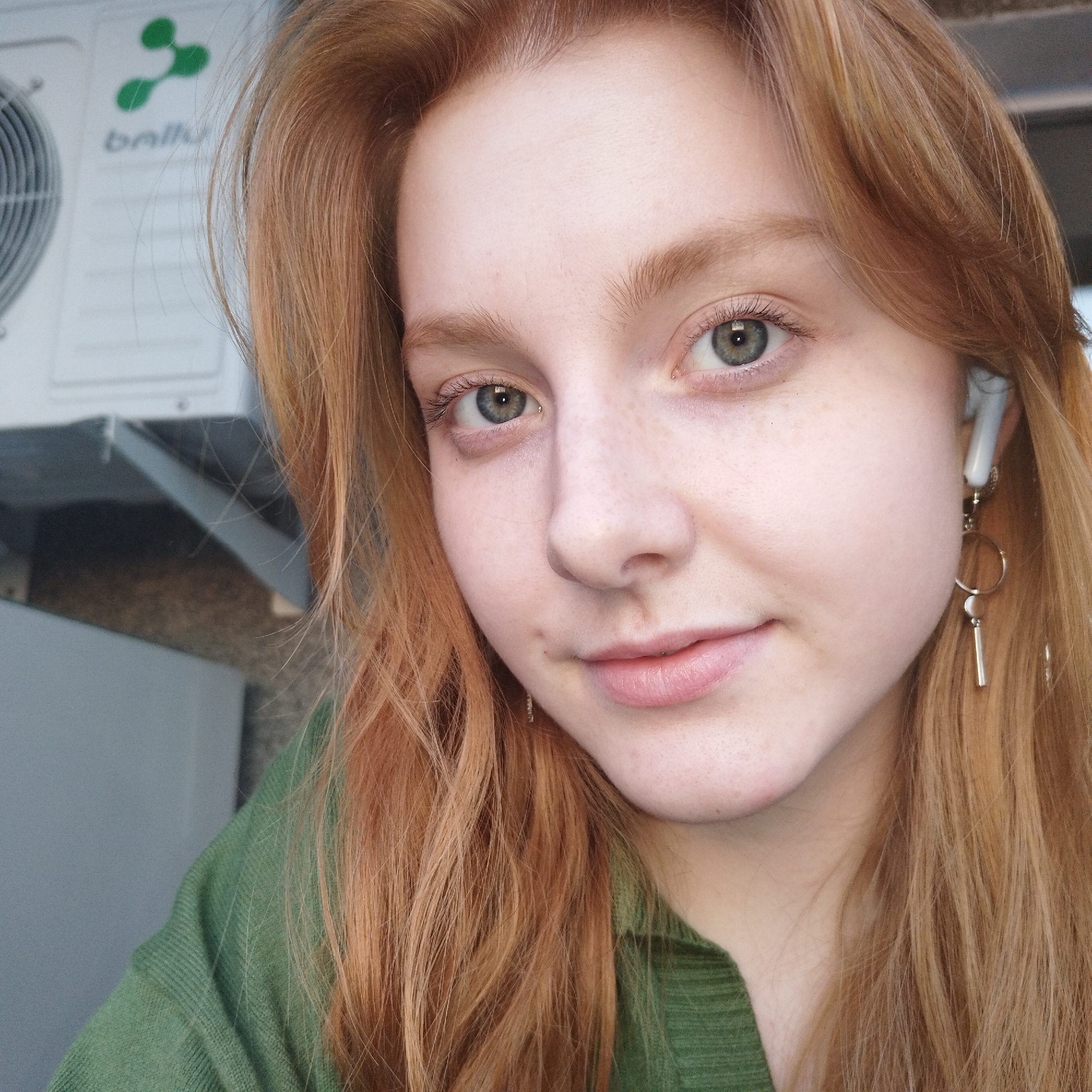 A close-up portrait features a young adult female with fair skin, light green eyes, and long, reddish-blonde hair. She exhibits freckles across her nose and cheeks, and her lips form a subtle smile. A white wireless earbud is visible in her right ear, alongside a metallic dangling earring composed of circular and bar elements. She is wearing a green, ribbed, long-sleeved garment. In the blurred background, an outdoor air conditioning unit, white in color, is visible to the upper left. The unit displays the "ballu" brand name and a green abstract logo, with a portion of its fan grille also discernable. The scene is stated by the user to be in Yekaterinburg, Russia.