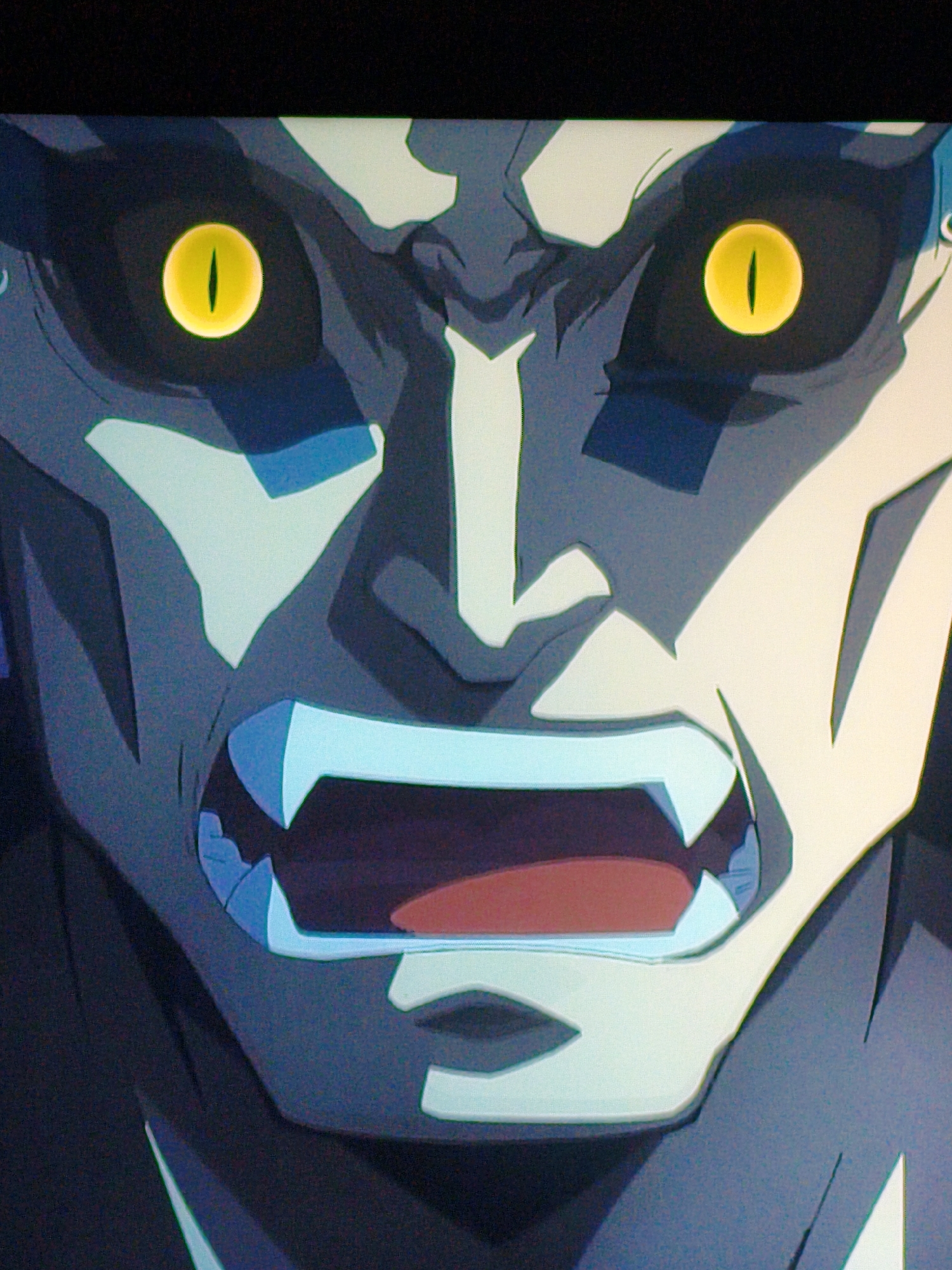 The image shows a close-up of an animated character's face, specifically its eyes, nose, and mouth. The character has striking yellow eyes with vertical pupils, giving it a fierce or monstrous appearance. Its mouth is open, revealing sharp, white teeth and a red interior. The character's skin is a dark gray with lighter, angular markings around its eyes and on its face. The animation style is distinct, with bold lines and sharp, geometric shapes. The background is dark, further emphasizing the character's features. There is no discernible location in Vladivostok, Russia, or any indication of a real-world environment.