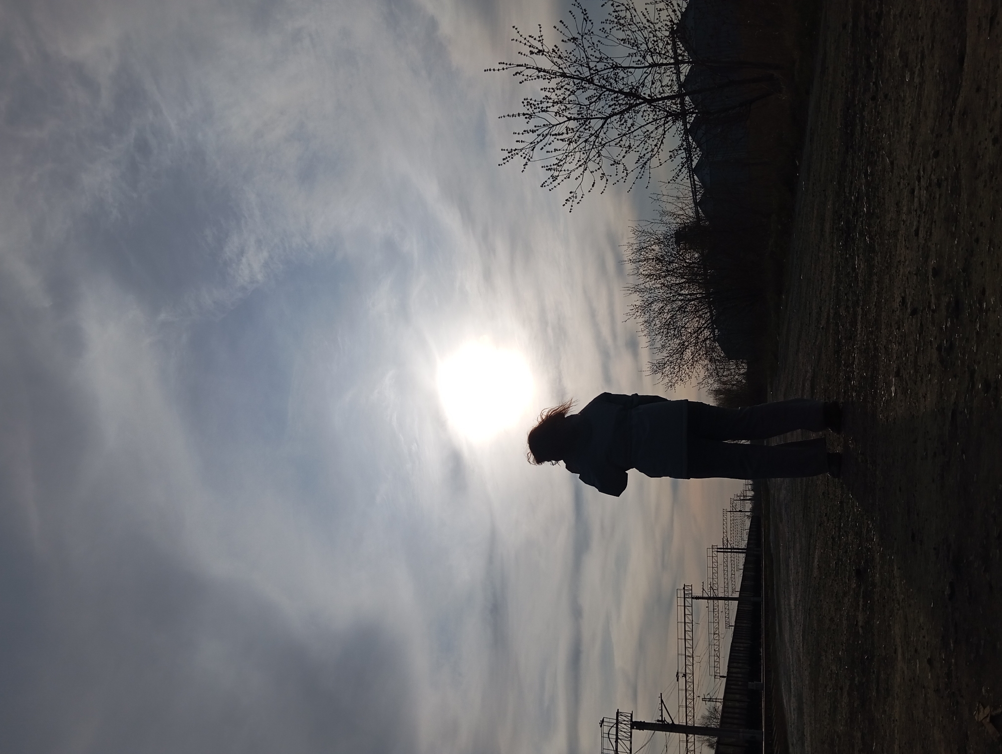 The image captures a striking silhouette scene, appearing to be taken either during sunrise or sunset, with the sun brightly positioned in the sky, directly behind the main subject. The overall composition is dominated by a strong contrast between the brilliant sky and the dark, silhouetted elements.
The primary subject is a lone person, seen from the side, standing on what appears to be a path or ground next to railway tracks. The person is completely silhouetted against the bright sky, making their features indistinguishable. They are wearing what looks like a jacket and trousers, and their hair, catching some light, appears to be flowing slightly, suggesting a breeze or movement. Their posture is somewhat bent forward, as if looking down or in deep thought.
The sky occupies a significant portion of the frame, filled with cirrus or altocumulus clouds that are stretched into long, wispy bands, reflecting the sunlight and creating a dramatic backdrop. The sun itself is a powerful white orb, casting a strong backlight.
In the midground and background, various elements are also rendered in silhouette. To the upper right, the bare branches of trees are visible, some with small buds, suggesting early spring or late autumn. Further along the horizon, more dense, dark masses of trees or industrial structures are present. Railway infrastructure, including poles and overhead lines, stretches into the distance on the lower left, indicating the setting is adjacent to train tracks. The ground where the person stands appears to be rough, possibly dirt or gravel.
The scene evokes a sense of solitude and contemplation, enhanced by the dramatic lighting and the silhouetted figure. Given the location context of Yurgamysh, Russia, it depicts a quiet moment in a possibly rural or semi-industrial landscape. There is no visible text in the image.