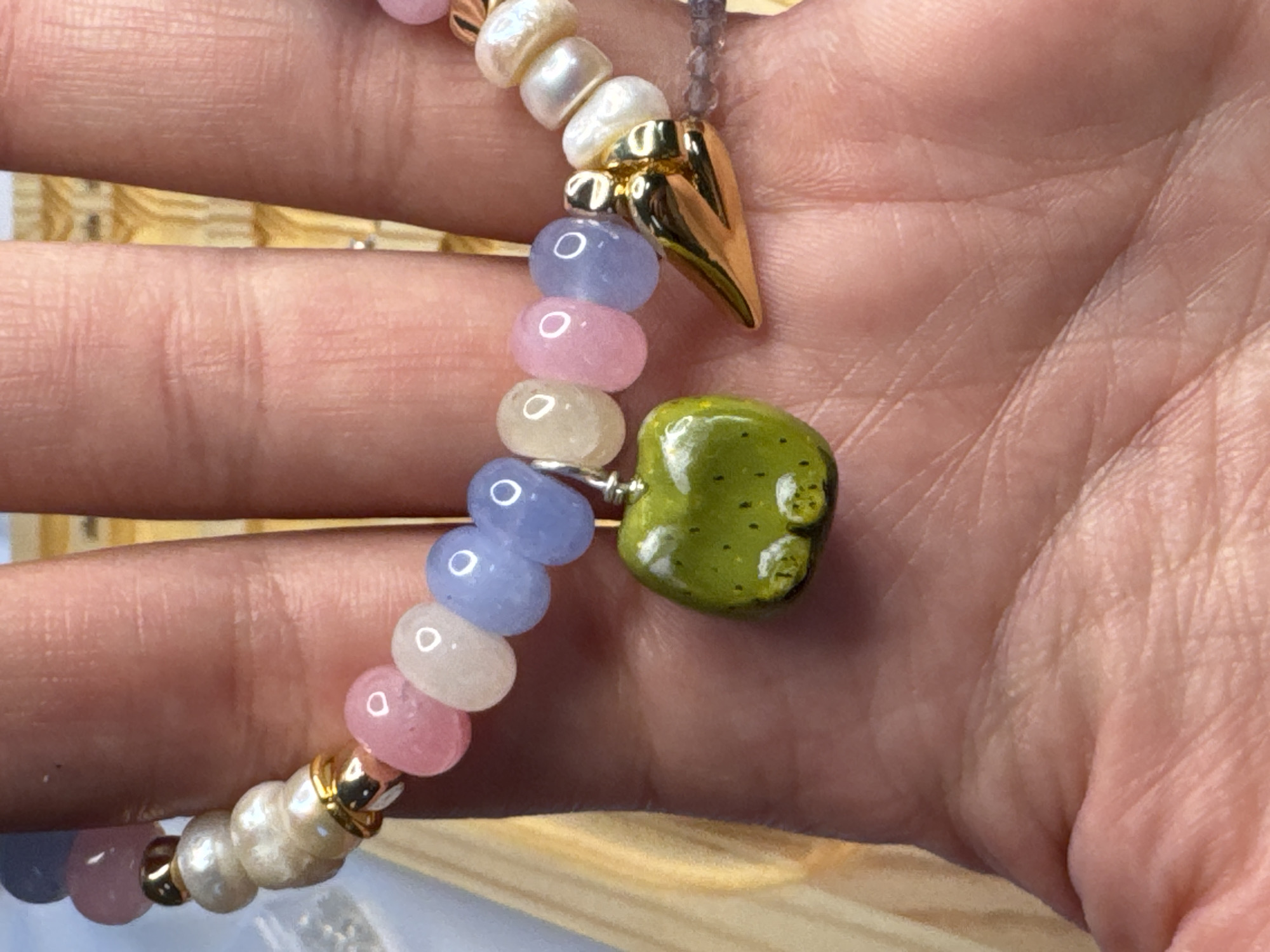 A close-up shot captures a hand adorned with a bracelet. The bracelet features a colorful assortment of beads, including pearls, pink, and blue stones, along with a charming green apple charm. A golden charm, shaped like a tooth, is also visible on the bracelet. The background appears to be a wooden surface, possibly a table or wall. The image does not provide any information about the city of Nikulino, Russia, or any specific location.