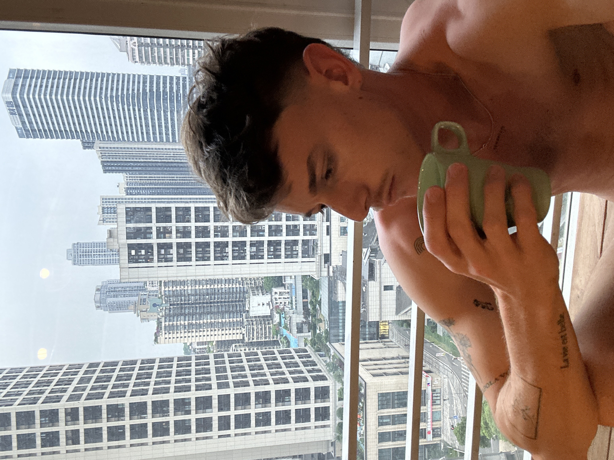 A shirtless man is seated indoors, holding a green mug near his face. He has a dark, cropped haircut, fair skin, and a necklace. Tattoos are visible on his forearms. Behind him, through a window, a cityscape is visible, comprising tall, modern buildings. A multilane highway is visible in the lower-right quadrant of the scene. The buildings suggest an urban environment, specifically the Yuzhong District in China.