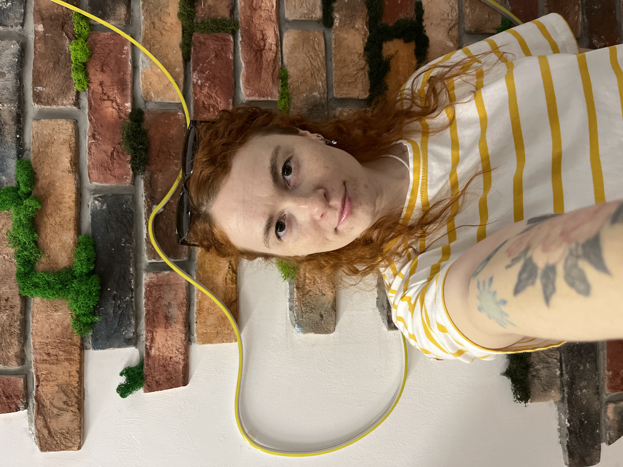 A selfie of a red-haired person wearing a white and yellow striped shirt. The person is positioned against a wall decorated with artificial brick and green moss. A yellow cable and sunglasses are visible in the background. The location is Chaikovsky, Russia.