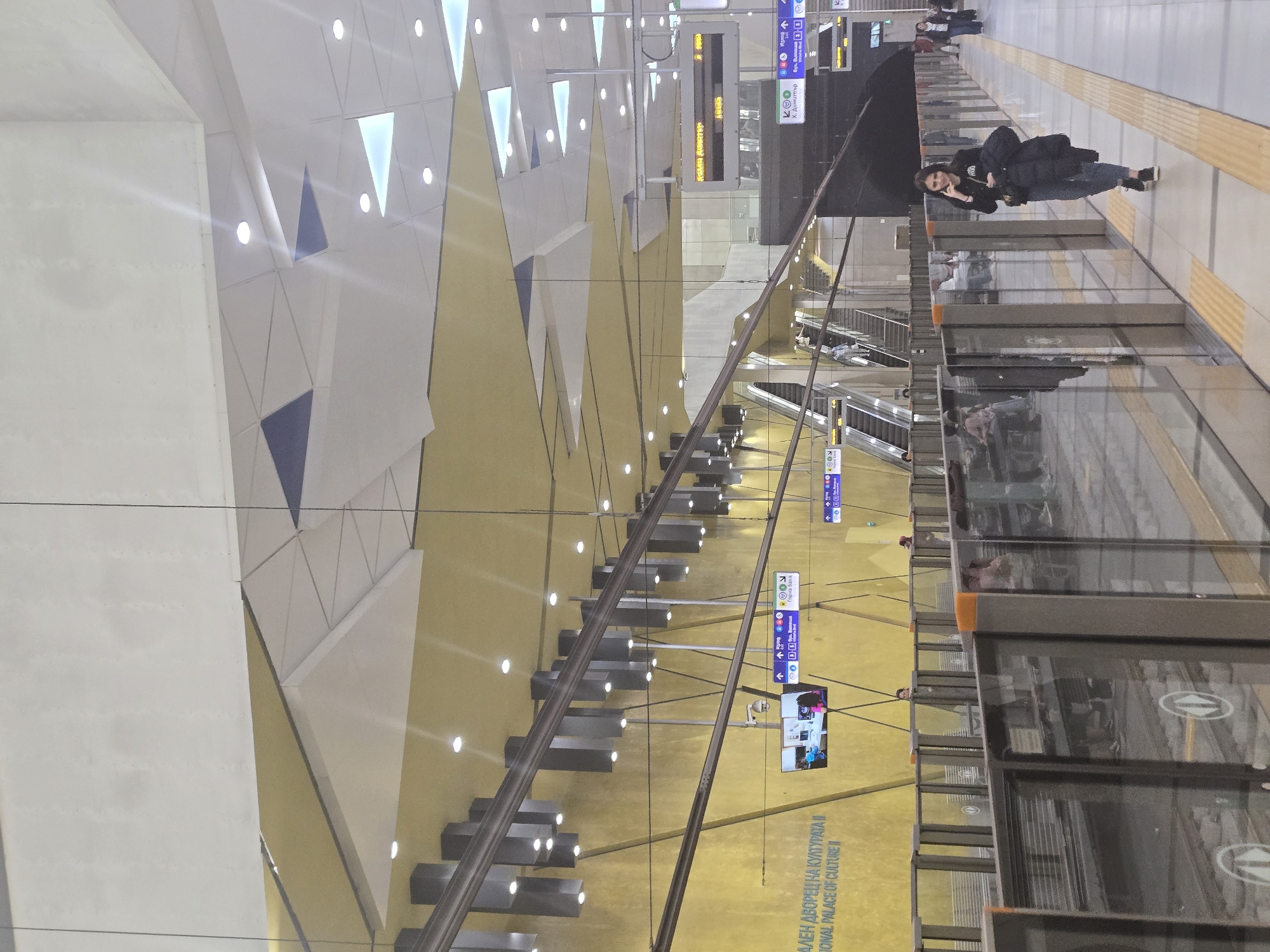 Here is a description of the image:
The image shows the platform level of a modern metro station in Sofia, Bulgaria. The station features a high ceiling with a geometric pattern of white triangles and angular shapes, accented with integrated lighting.
Along the platform edge, there is a safety barrier system with transparent panels and orange vertical supports. The floor is composed of light-colored tiles with a textured yellow safety strip along the edge.
A woman is visible on the platform, wearing a dark jacket and dark pants and looking at her mobile phone. The presence of station signage in Bulgarian indicates the location is Sofia, Bulgaria. The text on the signage in the image includes "АЛЕН ДВОРЕЦ НА КУЛТУРАΤΑ II," "Горна баня," and "Х. Димитър." This points to a station such as "National Palace of Culture II (NDK 2)" in Sofia's metro system.