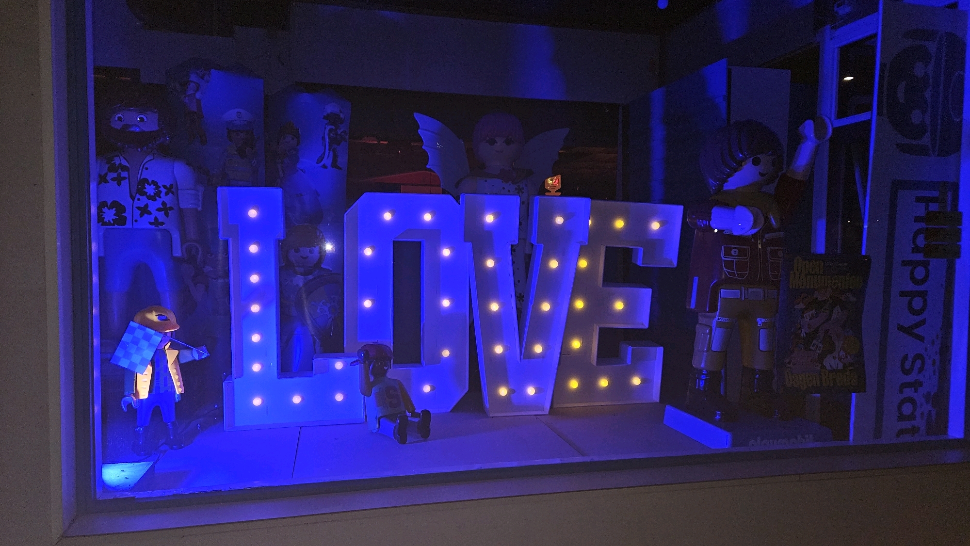 The image depicts a shop window display in Breda, Netherlands, featuring large illuminated letters spelling out "LOVE". The letters are white and studded with yellow lights. Several Playmobil figures are arranged within the display. On the left, a figure wearing a floral shirt and blue pants stands near the "L". To the right of the "L", a figure with wings, resembling an angel, is positioned behind the letters. Between the "O" and "V", a small Playmobil figure is seated. To the right of the "V", a larger Playmobil figure dressed in a red jacket and yellow pants stands with its arms extended. Behind this figure, a poster is visible with text that appears to include "Open Monumenten Dagen Breda". A vertical sign with the text "Happy stat" is on the far right. The entire scene is bathed in a strong blue light, giving it an artificial glow.