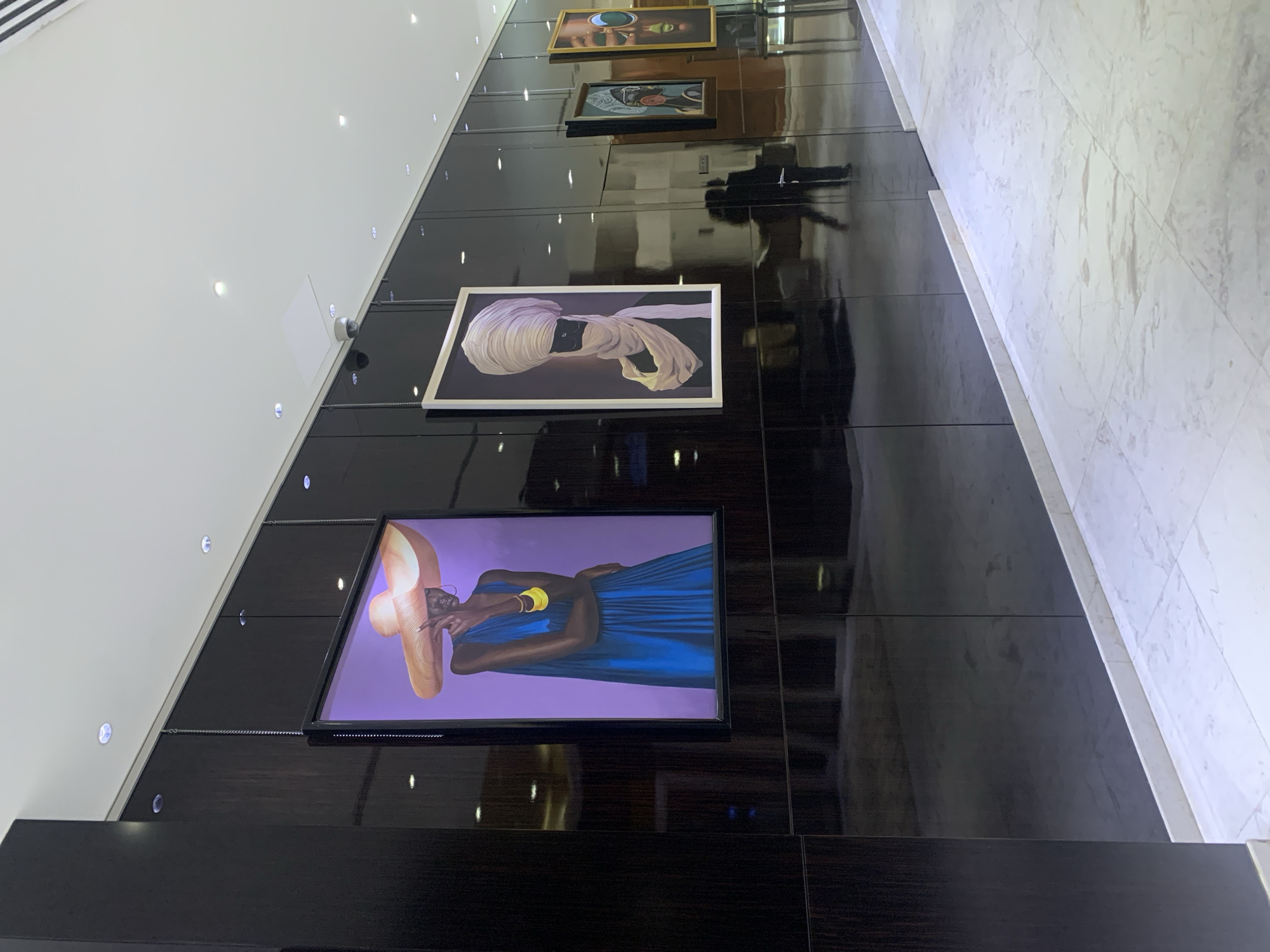 The photograph shows a corridor with artwork displayed on a dark-paneled wall. The floor is made of light-colored marble tiles. There are several framed artworks on the wall, including a portrait of a person wearing a large hat and a blue dress, and a second piece of art featuring someone wearing a white turban. The panels reflect the light from the wall and paintings. There are security cameras mounted near the ceiling and lights. The specific location within Dar es-Salaam, Tanzania, is not identifiable from the image alone.