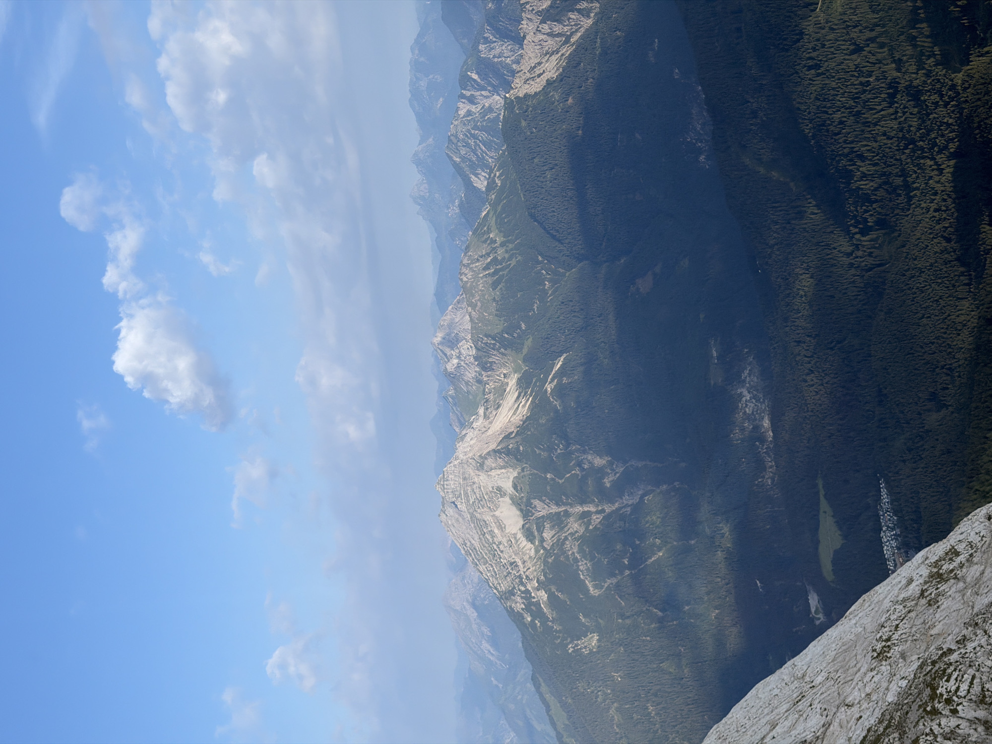 This image is a wide-angle, high-angle shot of a mountainous landscape near Grainau, Germany. The sky is a clear blue with scattered white clouds. In the foreground, a steep, rocky slope dominates the left side of the frame. The midground features several layers of forested mountains, with a prominent, rugged peak in the center, its upper slopes showing exposed rock and lighter vegetation. The background shows a series of hazy, blue-tinted mountain ranges receding into the distance. Nestled in a valley among the trees in the lower portion of the image, there is a small cluster of buildings, likely a village or settlement, characteristic of Grainau's alpine setting. The overall scene depicts a vast, natural environment with signs of human habitation subtly integrated into the landscape.