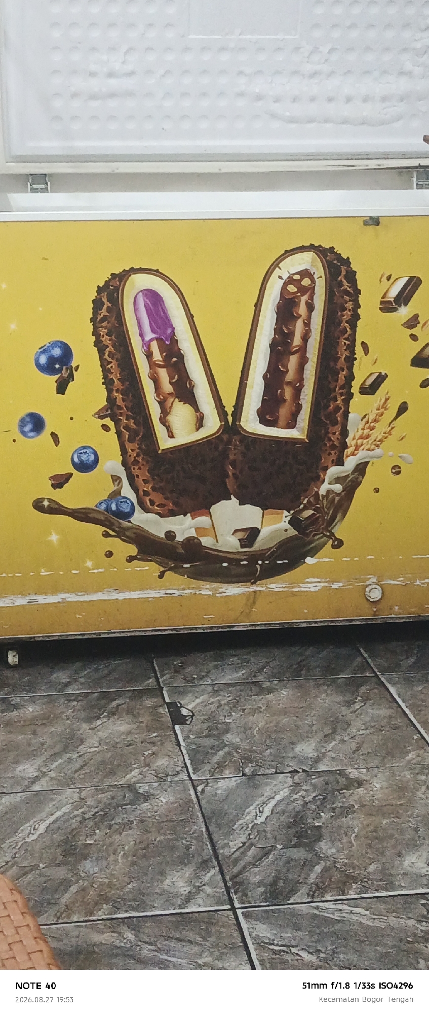 The image shows a close-up of a yellow refrigerator with an advertisement for ice cream. The advertisement features two ice cream bars with a bite taken out of each, revealing a purple filling in one and a chocolate filling in the other. Blueberries and chocolate pieces are scattered around the ice cream bars, along with splashes of chocolate sauce and what appears to be wheat stalks. The bottom of the image shows a tiled floor. The text at the bottom indicates the location is "Kecamatan Bogor Tengah," which is a subdistrict in Bogor, Indonesia, and the date and time are "2026.08.27 19:53." The camera settings are also listed as "51mm f/1.8 1/33s ISO4296."