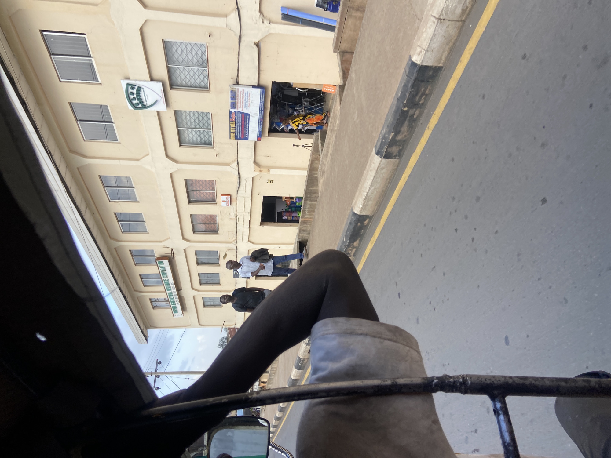 The image captures a street scene in Jos, Nigeria, as seen from the perspective of someone in a motorized rickshaw. The building in the background is a light-colored, multi-story structure with numerous windows. One prominent sign on the building reads "NIGERIAN AGRICULTURAL INSURANCE CORPORATION". Another sign, partially obscured, appears to advertise "RENTAL CARS". Several shops are visible at street level, one with an open doorway revealing products inside, and a person in bright yellow and orange clothing standing within.
Two men are standing on the sidewalk in front of the building. One is wearing a white shirt and blue jeans, holding a jacket over his shoulder. The other is wearing a dark t-shirt. The street in the foreground has a yellow line marking the edge of the road and a curb with black and white stripes. The sky is overcast, suggesting it might be a cloudy day. The lighting is diffused, indicating it is likely daytime, possibly mid-morning or afternoon. The perspective suggests the rickshaw is moving along the street. The overall scene depicts a typical urban environment with commercial activity.