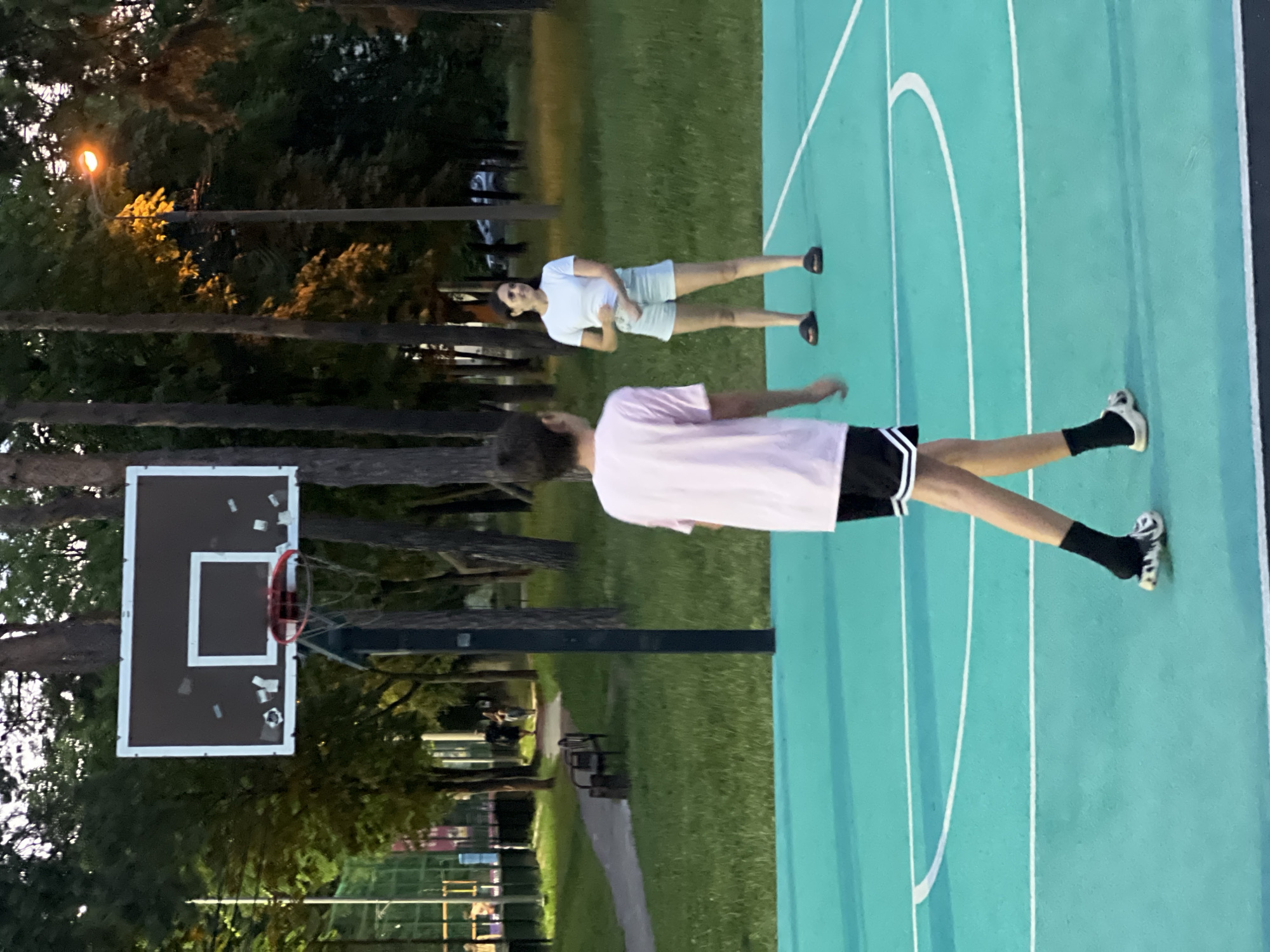 The image depicts an outdoor basketball court, likely in Krasnodar, Russia, given the context. Two individuals are present on the court: a young person in the foreground wearing a light pink t-shirt, black shorts with white stripes, black socks, and white and black patterned shoes, and a woman in the middle ground wearing a white t-shirt and light blue shorts, with black footwear. The woman is looking towards the young person and appears to be smiling. The basketball court has a bright turquoise surface with white boundary lines. In the background, trees and grass are visible, with a lamppost casting an orange glow. A basketball hoop with a backboard is positioned on the left side of the frame. The lighting suggests it is either late afternoon or early evening.