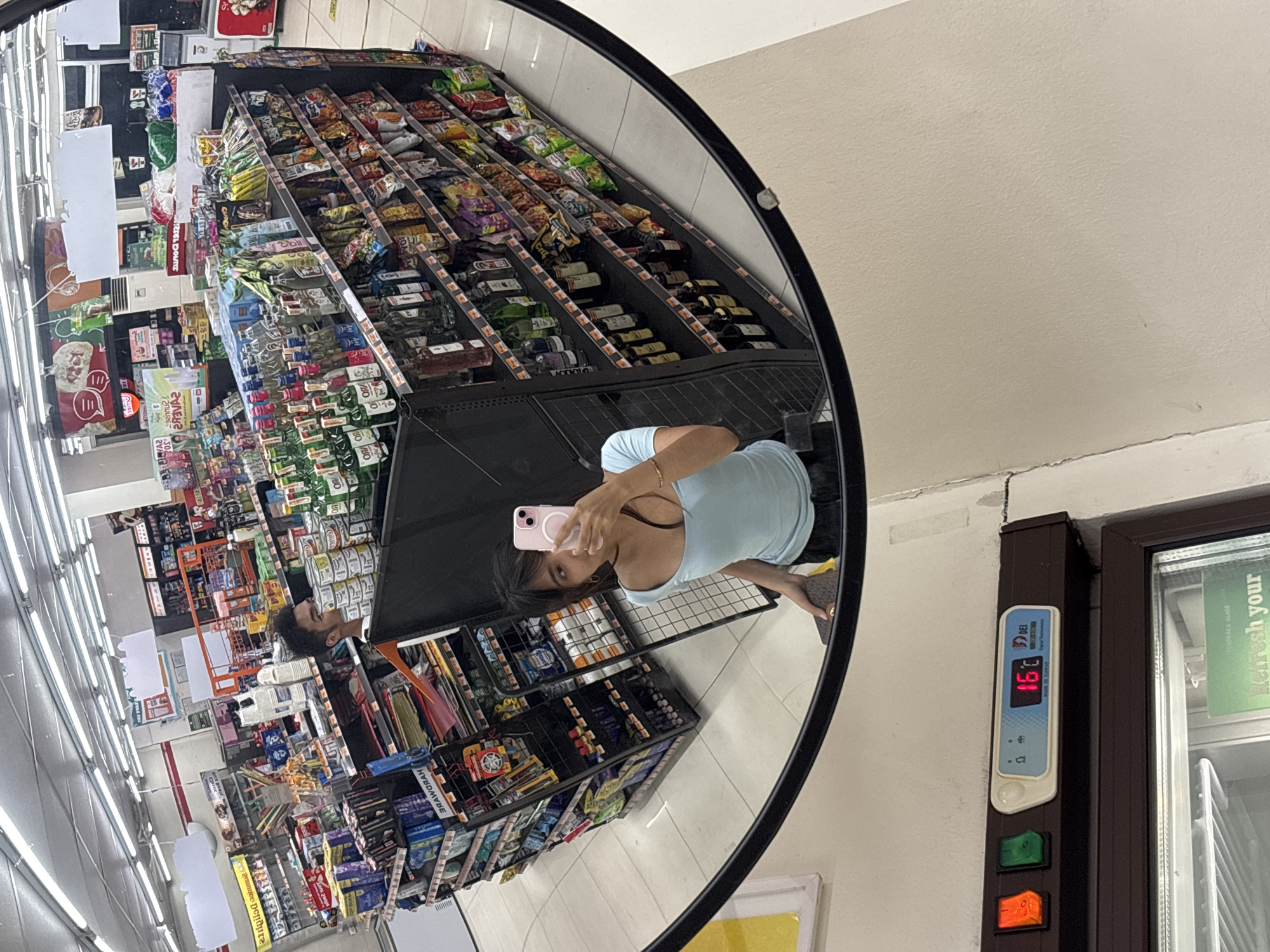 The image shows a mirror reflection of a woman taking a selfie in what appears to be a convenience store. The store is stocked with various items, including bottled beverages, snacks, and what seems to be canned goods. In the background, several aisles of products are visible, with fluorescent lighting illuminating the space. A man can be seen in the distance, partially obscured by a shelf. A digital thermometer on a refrigerator unit shows a temperature of 16 degrees Celsius. The overall environment suggests an indoor retail setting, likely a small convenience store or a similar establishment. Based on the presence of a DEI Digital Thermometer with "Made in Taiwan" printed on it, and general store aesthetics, this could be a 7-Eleven or a similar franchise store located in Davao City, Philippines.