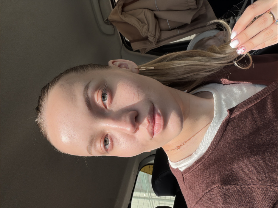 The image displays a close-up portrait of an adult female individual seated inside a vehicle. The subject has fair skin, light brown hair pulled back into a ponytail, and green-blue eyes. She is wearing a white top beneath a dark brown v-neck cardigan. Her lips appear to have a glossy finish. Accessories include gold-toned chain-link earrings on her right ear (viewer's left), a delicate gold-toned necklace with a small heart-shaped charm, and a ring with a light blue stone on her left ring finger (viewer's right). Her left hand is partially raised, displaying manicured nails painted white with small pink floral or dot patterns on the ring and pinky fingers. Bright natural light illuminates the scene from the left, casting shadows across the subject's face. The background reveals an automobile interior with a dark headliner, a dark car seat visible to the left, and a light-colored jacket hanging on a hook on the right side of the vehicle's cabin. A blurred exterior view is visible through the window on the left. The specific location within the city of Bezborodovo, Russia, cannot be determined from the visual information provided.