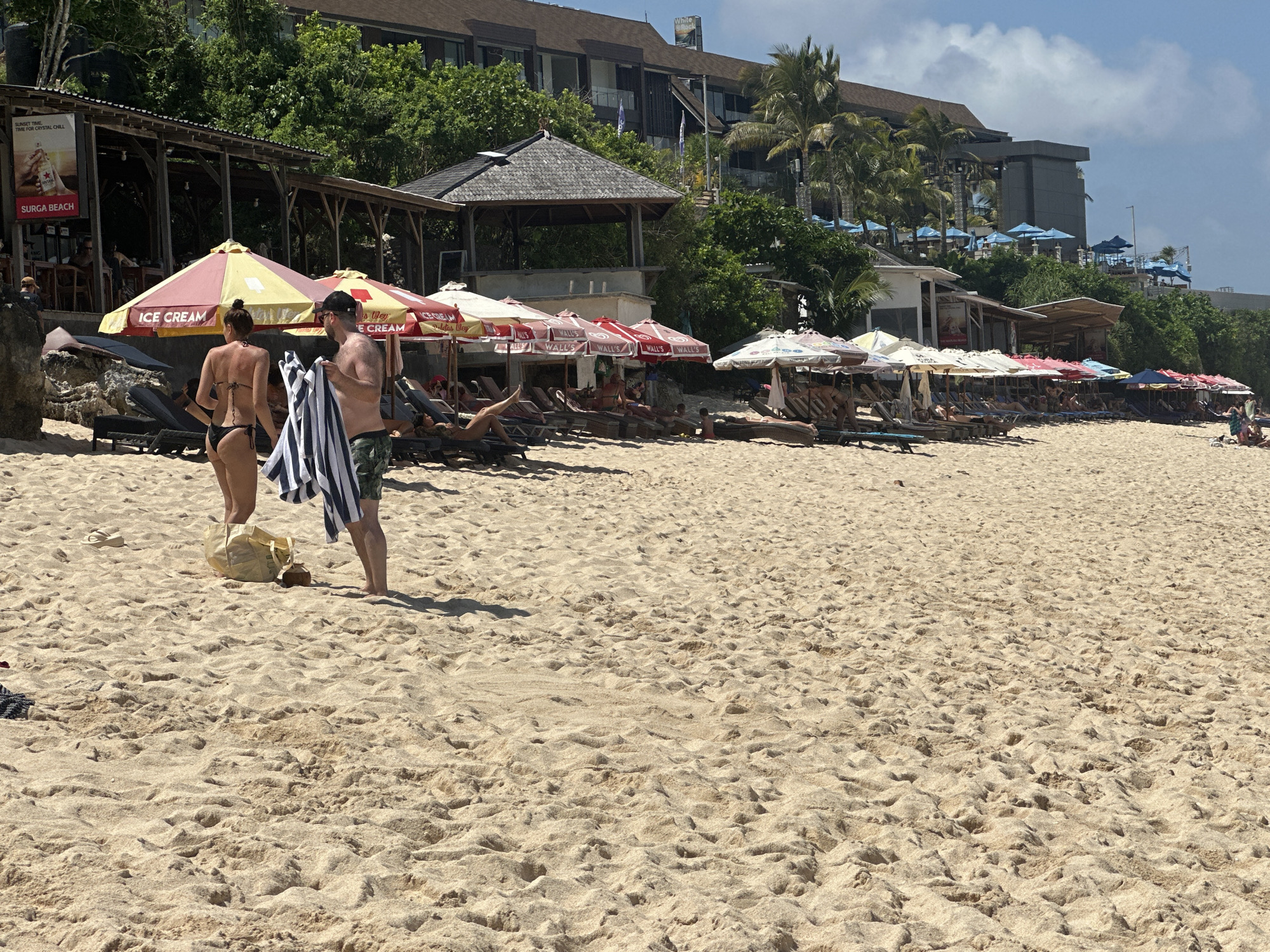 This is an outdoor shot of a beach in Kuta Selatan, Indonesia, densely populated with people relaxing in lounge chairs under a series of colorful beach umbrellas. A couple stands in the foreground, preparing for a day at the beach. The woman wears a bikini, while the man holds a striped towel. A beach bag and coconut drink sit on the sand near them. In the background, there are several open-air structures, one a restaurant bar, and a multi-story hotel structure sits atop the hill overlooking the beach. The sky is light blue with scattered clouds, suggesting a sunny day.