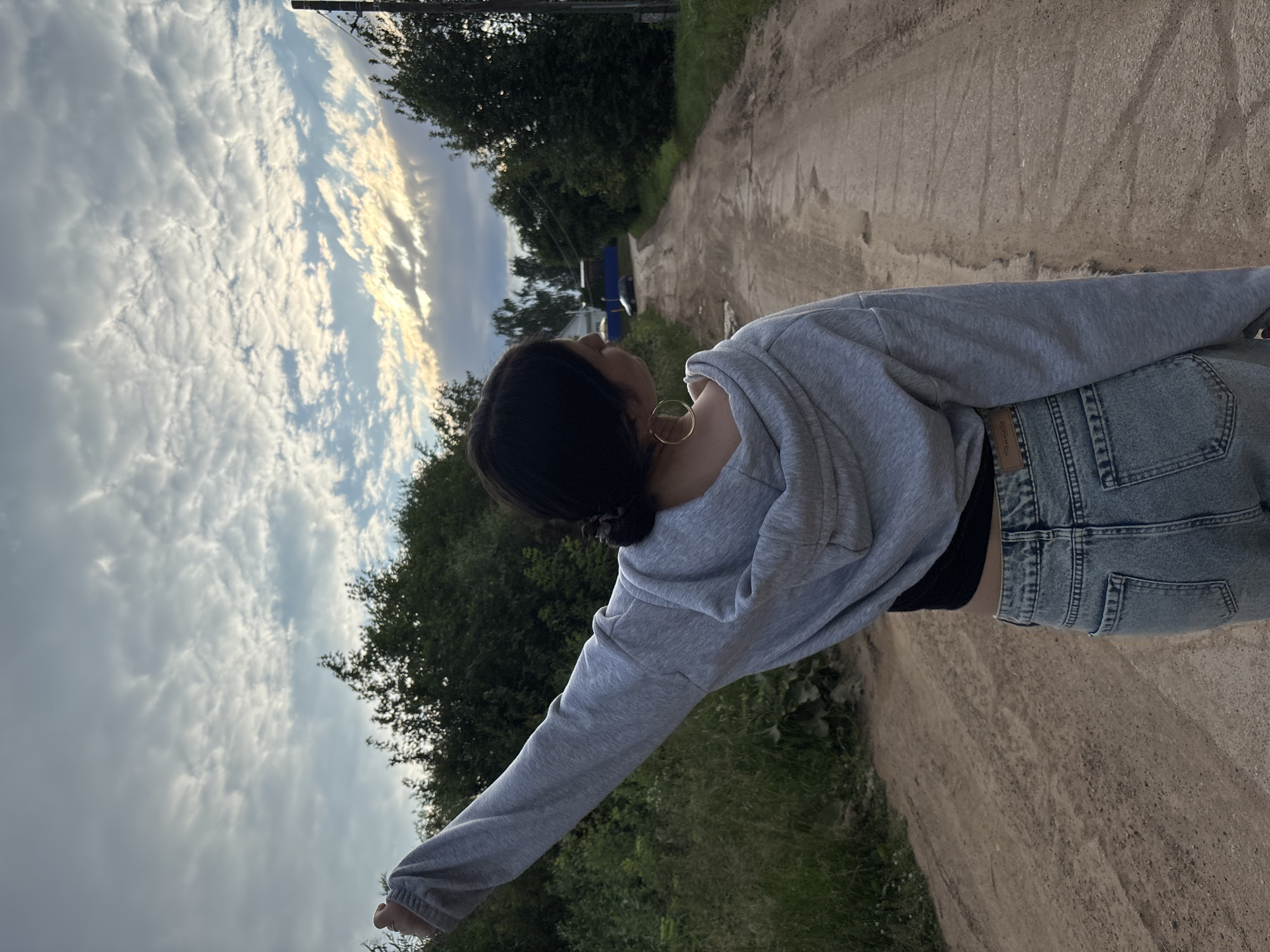 The image depicts a young woman standing on a dirt road in the city of Fokino, Russia, with her back to the camera. She is wearing a light grey hooded sweatshirt, a black crop top, and light-wash denim jeans. Her right arm is raised upwards and outwards. The road stretches into the distance, lined with trees on either side. The sky above is partly cloudy with streaks of sunlight breaking through. In the distance, a dark-colored car and a building are visible. The overall scene suggests an outdoor, rural setting.
