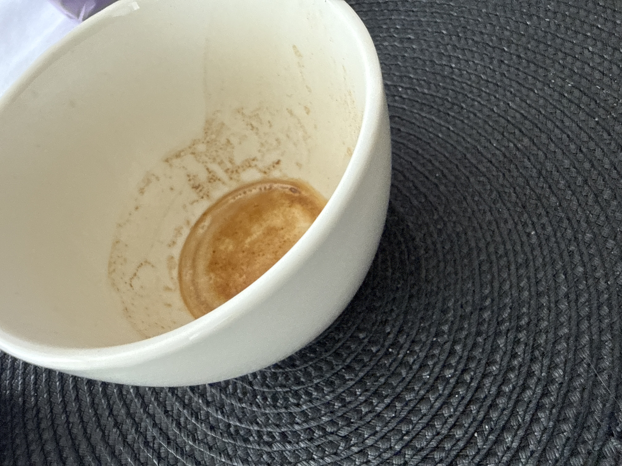 The image shows an empty, white ceramic coffee cup with residue from coffee on the inside, resting on a black, woven placemat. The placemat has a circular, textured pattern. The setting appears to be indoors, with a light-colored background visible in the upper right corner. There is no indication of the specific location within Burgas, Bulgaria.