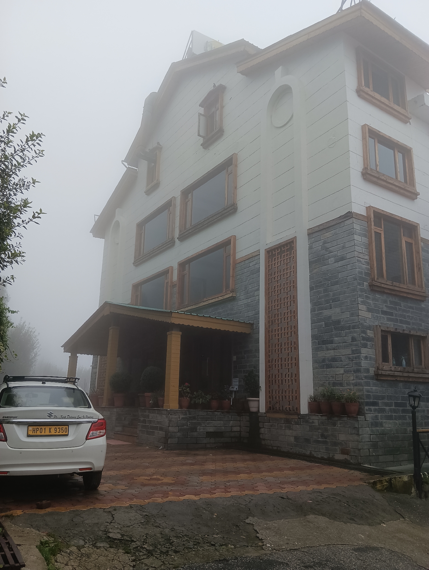 A white sedan with license plate HP01 K 9350 is parked on a patterned paved driveway in front of a multi-story building. The building has white and gray brick siding, numerous windows with wooden frames, and a covered entryway supported by wooden pillars. The scene is obscured by fog, with only the lower portion of trees visible on the left side. The location is Siumsa, India.