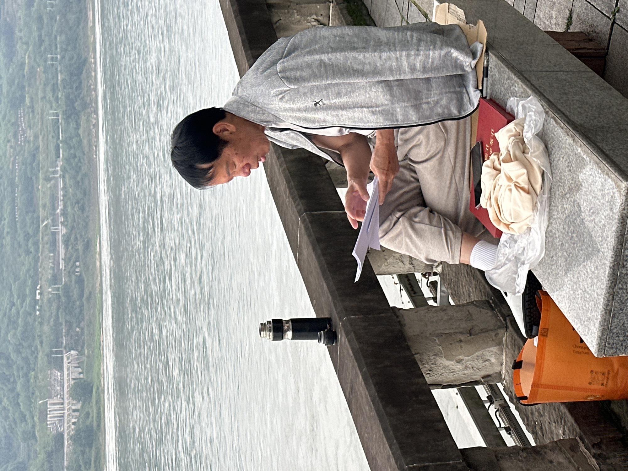 On a clear day, an Asian man is seated on the edge of a stone platform overlooking a vast body of water, likely the Yangtze River near Jiangjin, China, based on the surrounding landscape and architecture. The man is wearing a light gray jacket, light-colored pants, and white socks. He is holding a piece of paper in his hands, appearing to read or study it intently. To his right, on the platform, there is a red box with a white cloth or bag on top, suggesting he may be resting or working there. A small metal object or device is affixed to the railing in front of him. An orange shopping bag is on the platform beneath the man's feet. The environment features a landscape of trees and buildings across the river, typical of the region.